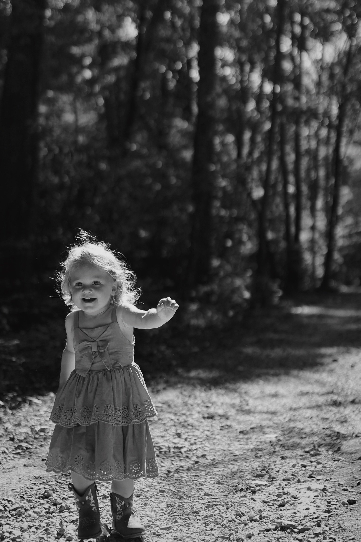 A young girl with curly hair, wearing a sleeveless dress with lace details and cowboy boots, is smiling and reaching out with her right hand while standing on a dirt path surrounded by trees in a forest, in black and white.