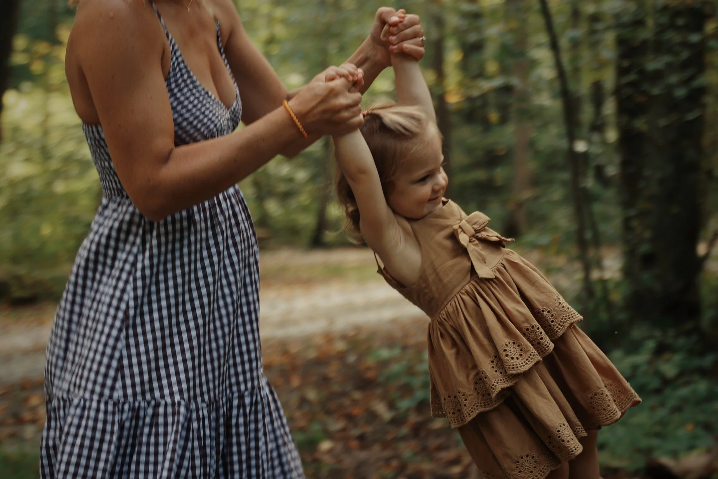A woman wearing a black and white checkered dress swinging a young girl in a tan dress with eyelet detailing, outdoors in a wooded area.
