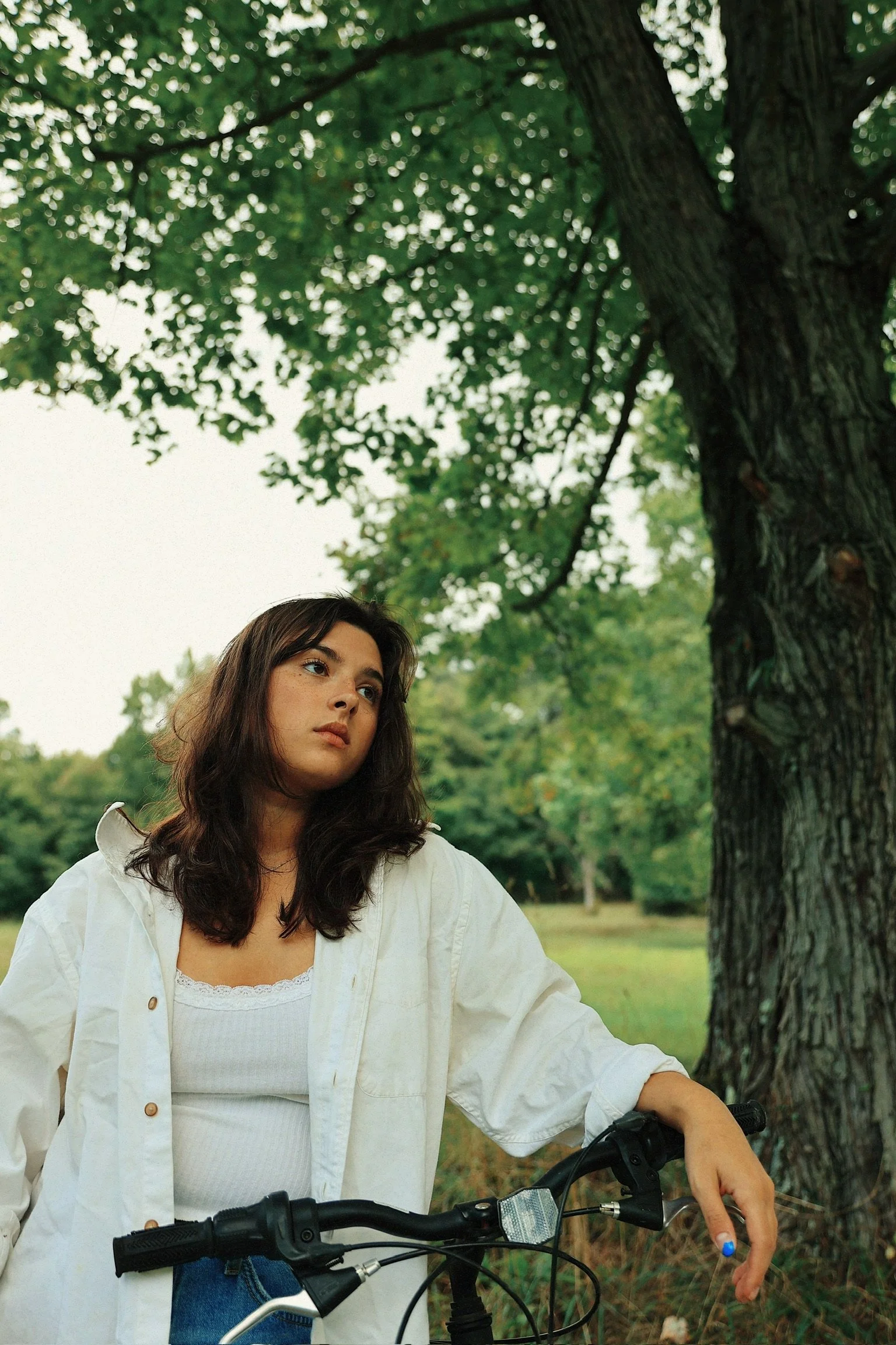 A young woman with long dark hair, wearing a white shirt and a white jacket, sits on a bicycle in a park with green trees and grass, looking thoughtfully into the distance.