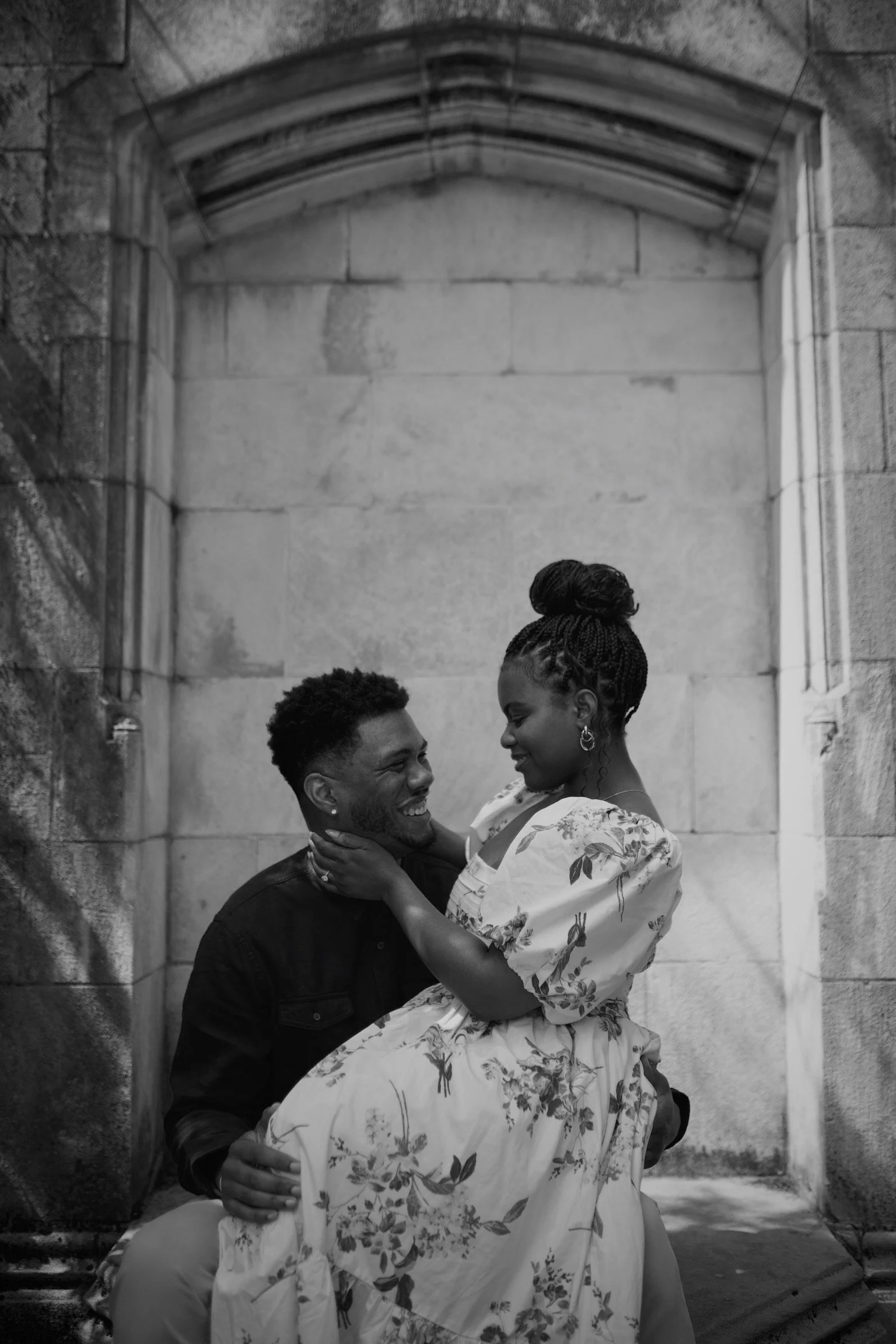 A black-and-white photo of a couple, with the man sitting and holding the woman, who is sitting on his lap, facing him and smiling. They are in front of a stone wall.