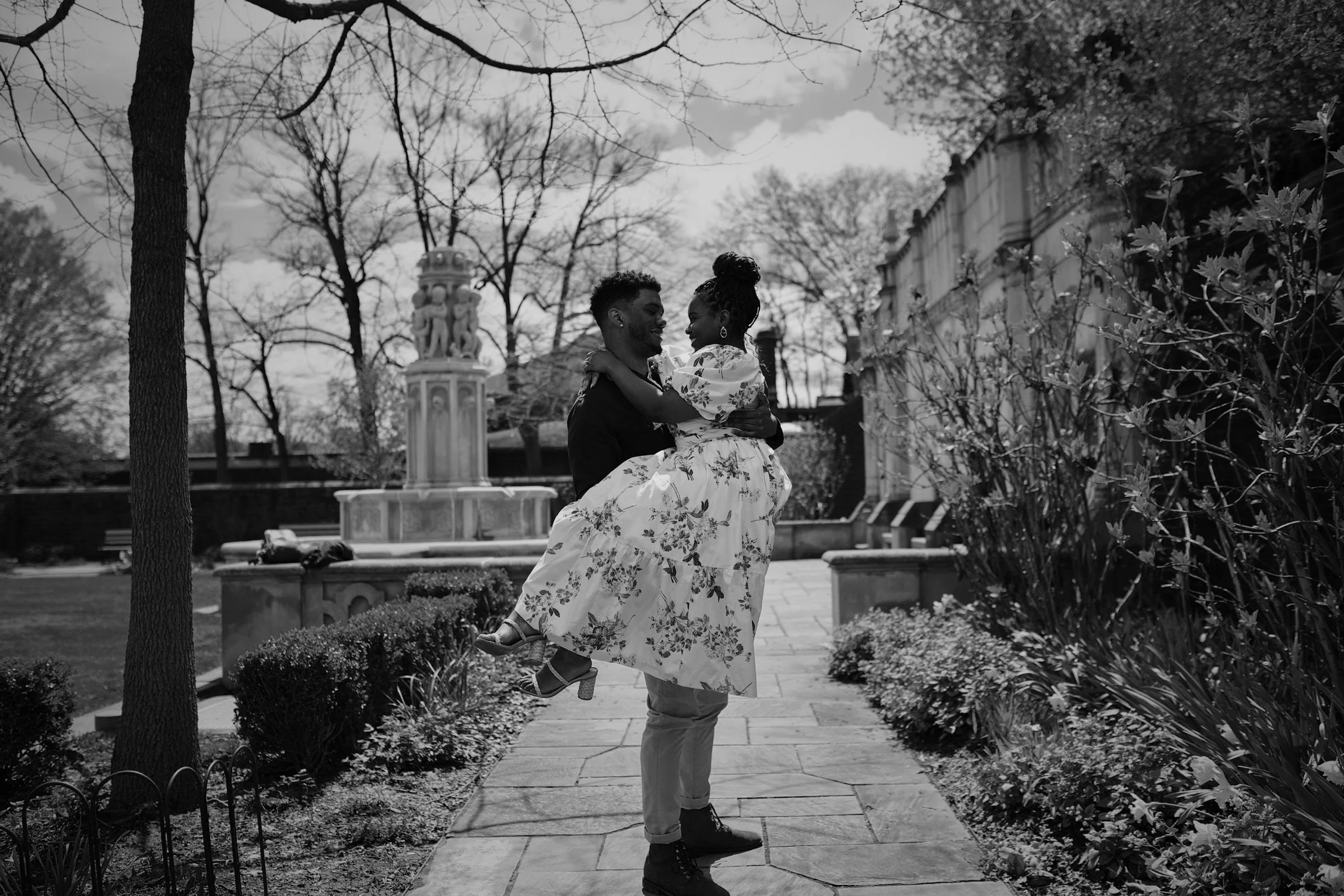 A man lifting a woman in a floral dress in a park with trees and a fountain in the background.