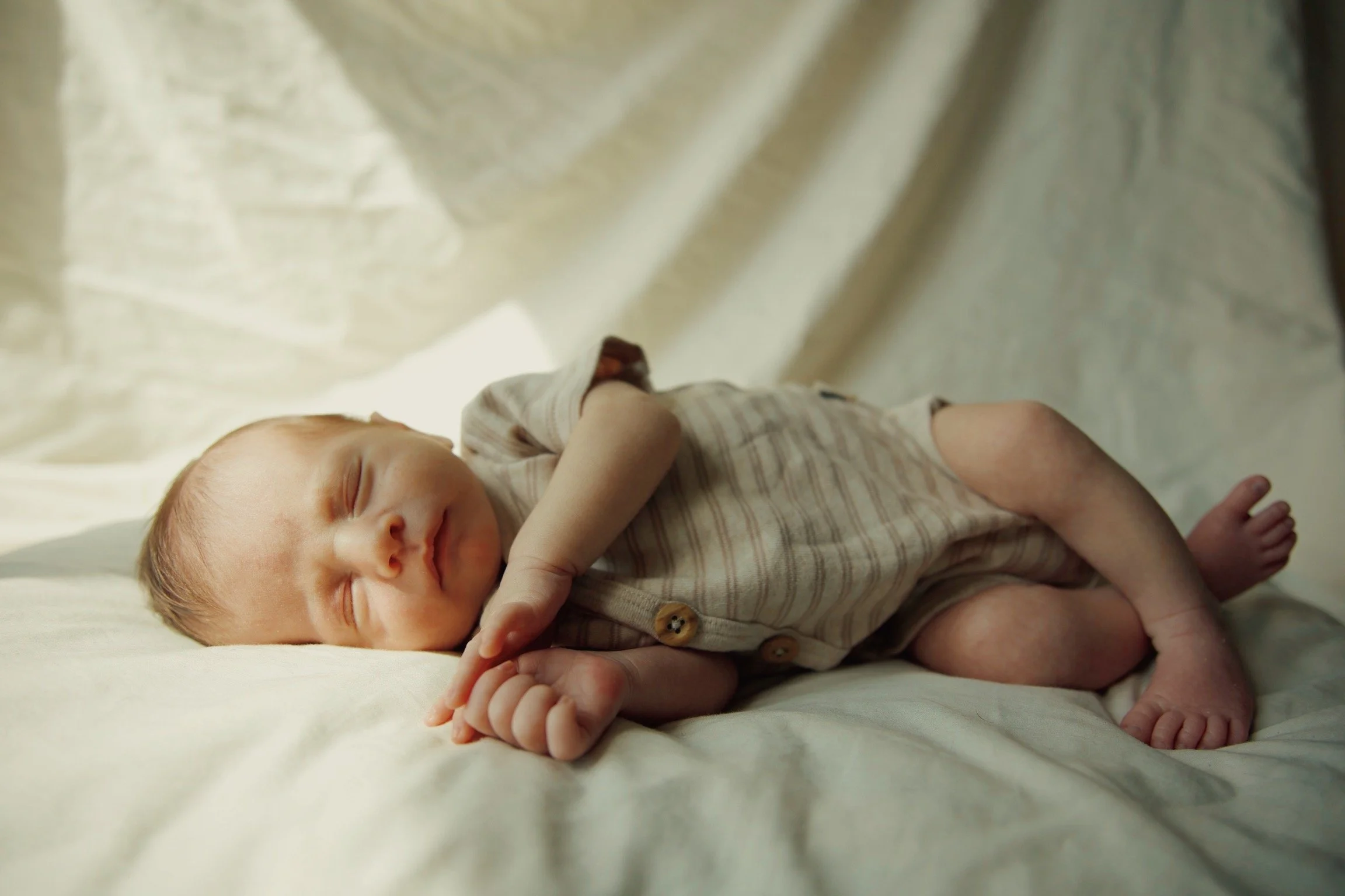 Baby lying on its side, peacefully sleeping on a soft, light-colored surface, wearing a beige and white striped onesie.
