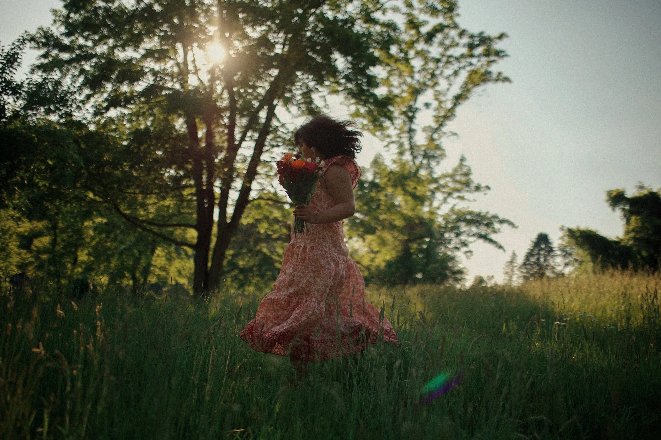 A woman in a flowing patterned pink dress holding a bouquet of flowers walking through a grassy field with trees in the background during sunset.