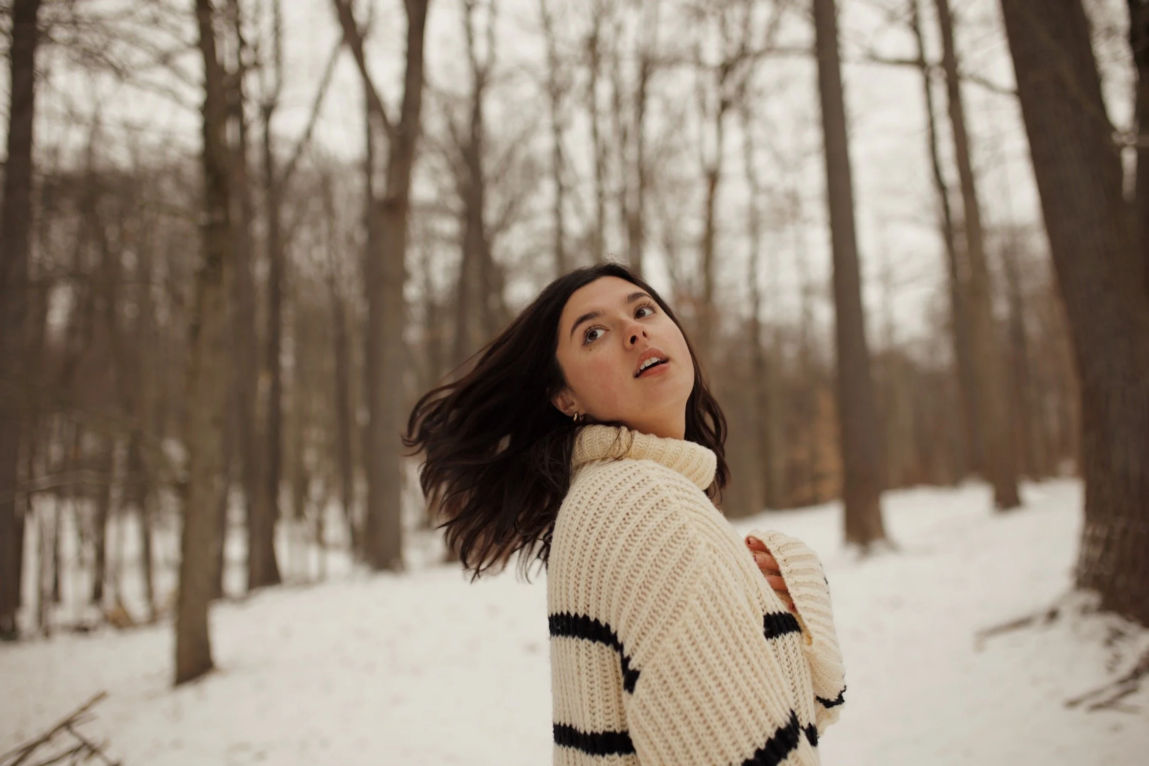A woman with dark hair wearing a cream-colored sweater with black stripes, standing in a snowy forest during winter, looking over her shoulder.