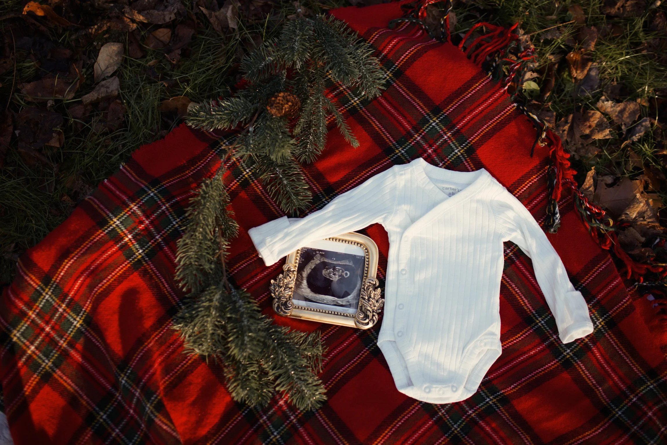 A white baby onesie on a red plaid blanket with pine branch decorations and a framed black-and-white ultrasound photo.