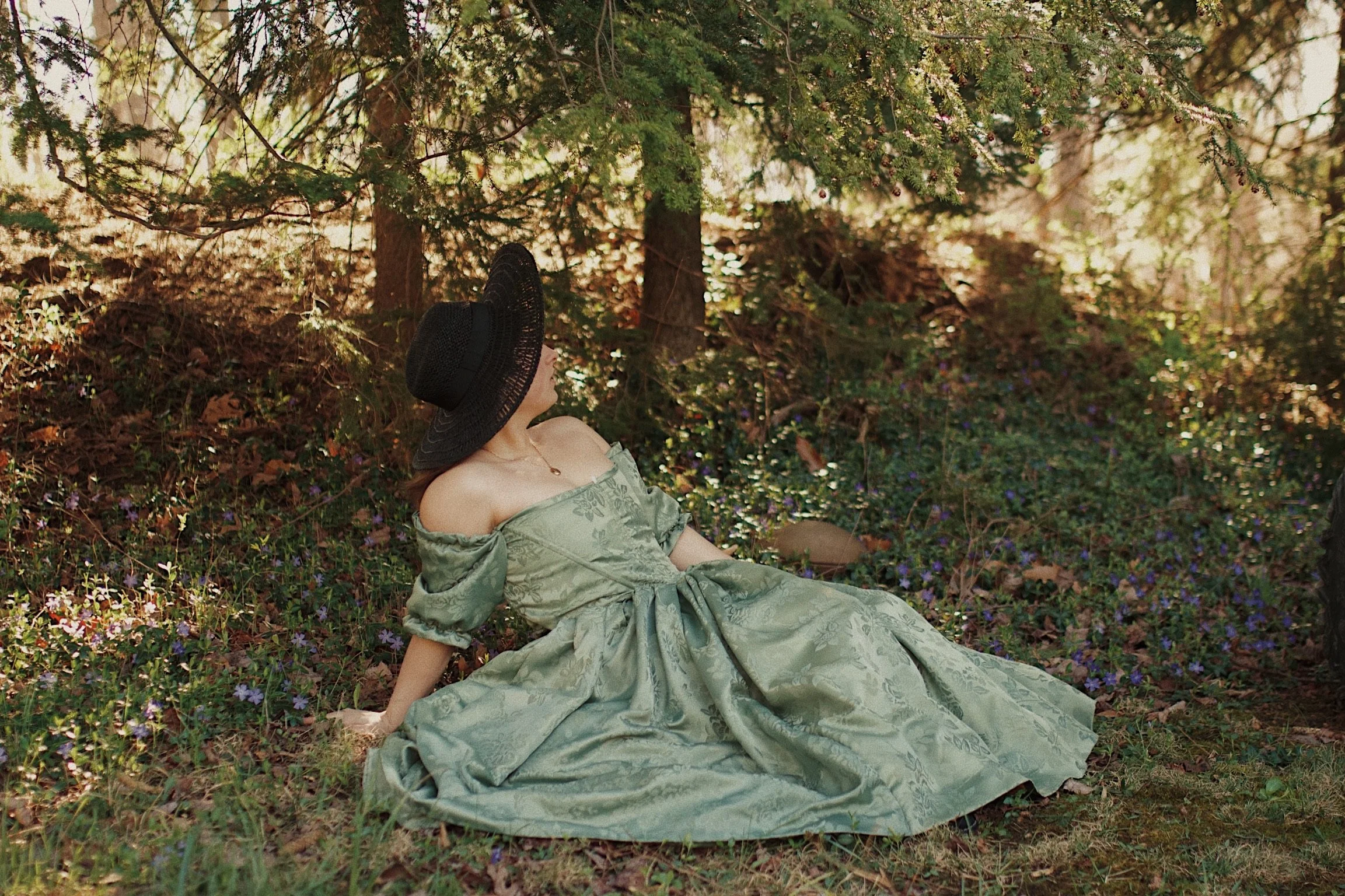 A woman wearing a green off-the-shoulder dress and a wide-brimmed black hat is sitting on the ground in a forest surrounded by green foliage and small purple flowers.