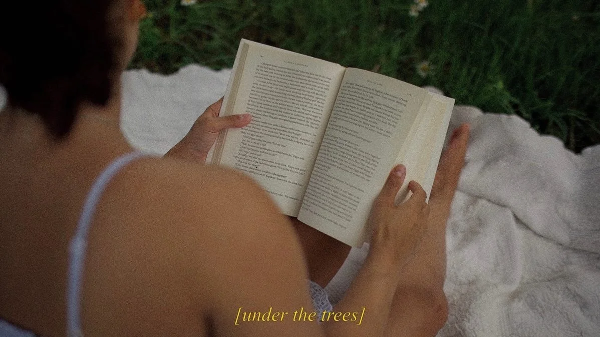 A woman sitting outdoors on a blanket, reading a book under a tree.