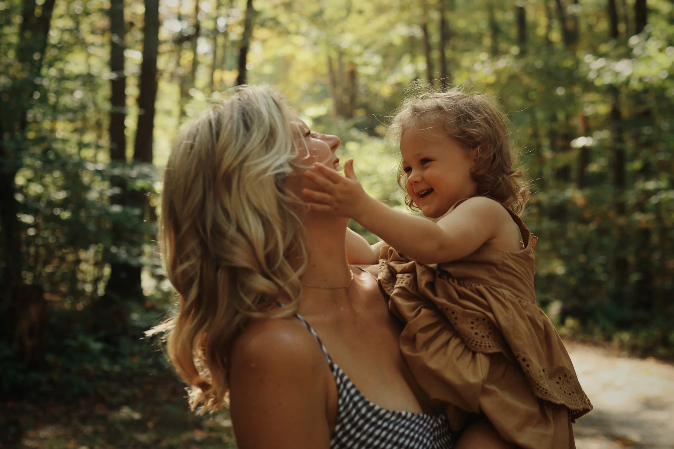 A woman and a young girl in a forest, smiling and playing together. The girl touches the woman's face softly.