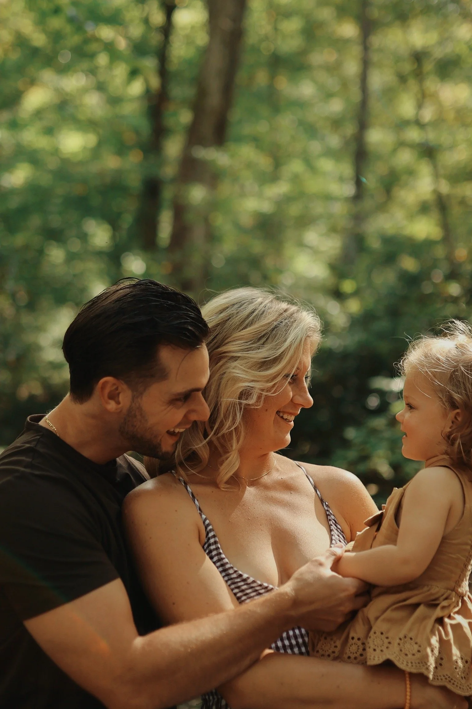 A family of three—mother, father, and young daughter—smiling and interacting outdoors in a lush, green forest.