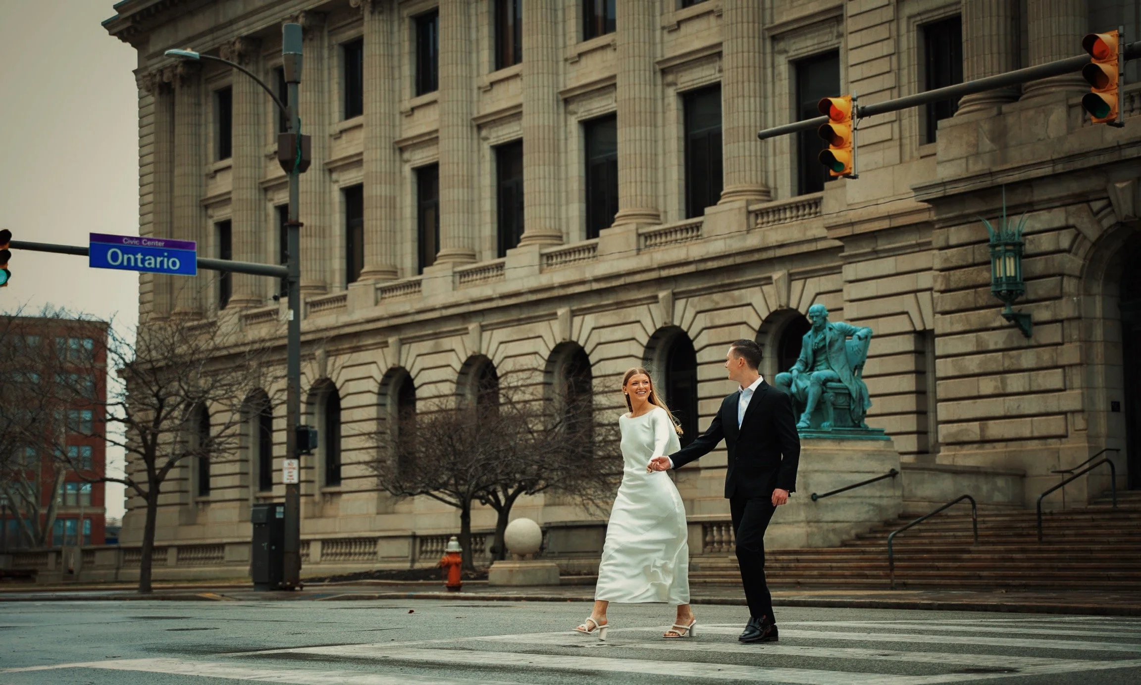 A couple dressed in formal attire walking hand-in-hand across a city street, with an ornate building and a bronze statue in the background.