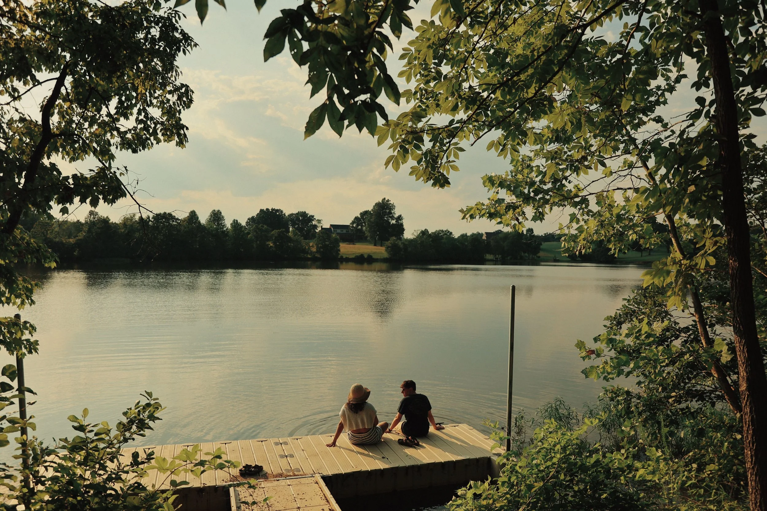 A woman and a man sitting on a dock by a lake, surrounded by trees and greenery, with a cloudy sky overhead.