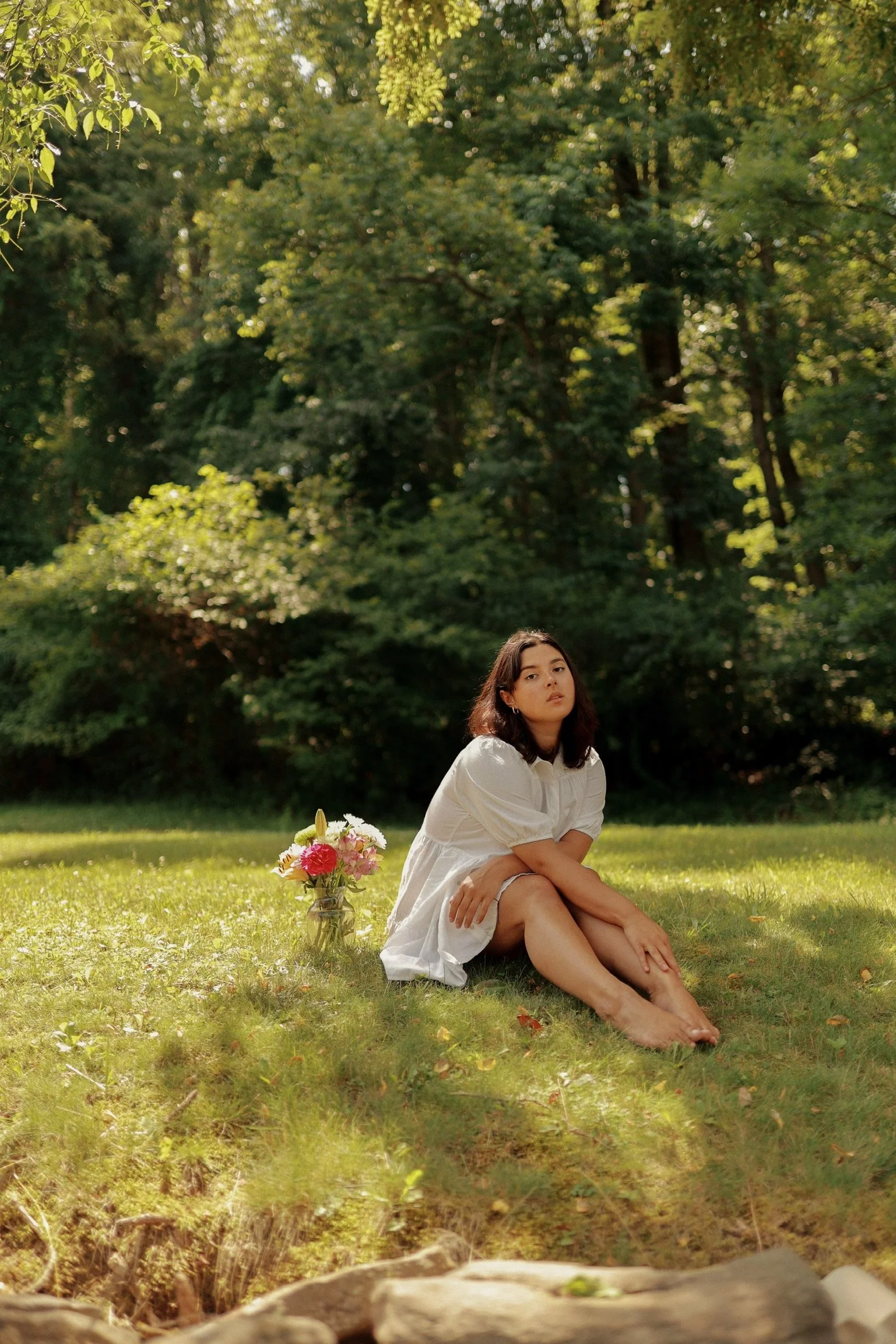 A young woman in a white dress sitting on the grass in a park or forested area, with a bouquet of flowers beside her, surrounded by lush green trees and sunlight filtering through the leaves.