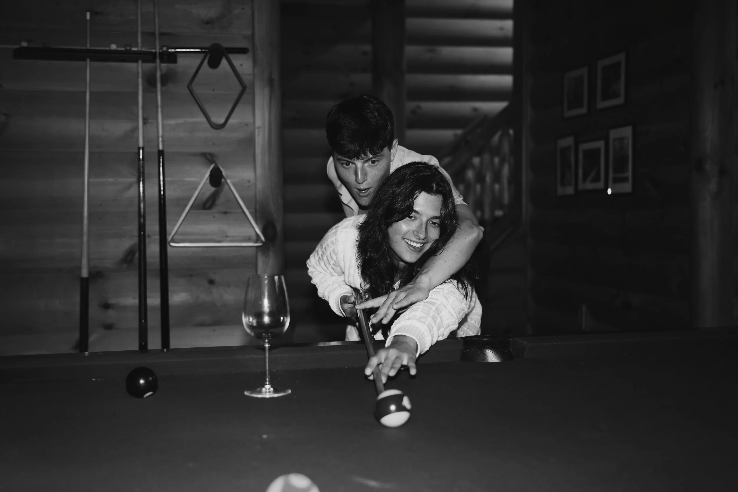 Two young adults playing pool, the woman is aiming to take a shot, the man is standing behind her with a surprised expression, in a cozy wooden room with framed pictures on the wall.