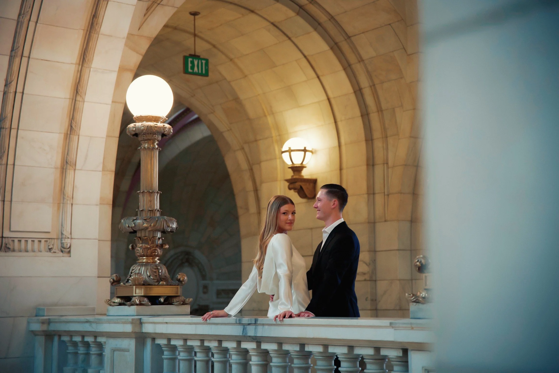 A couple dressed in formal attire standing on a marble balcony in a historic building, with arched stone walls and ornate lighting fixtures.
