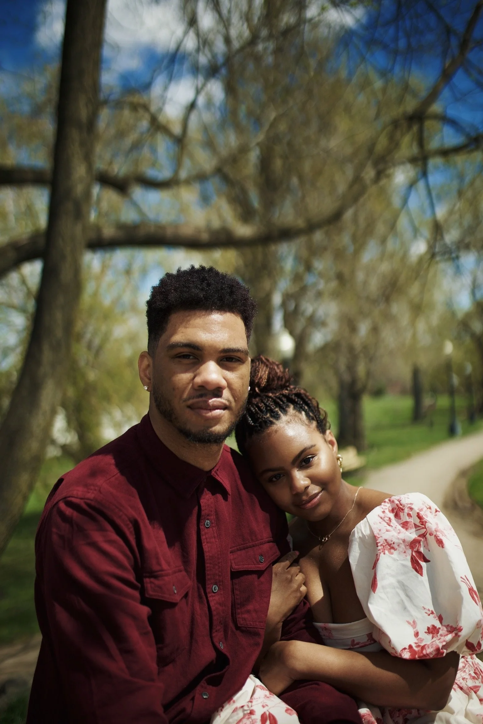 A young couple sitting close together outdoors on a sunny day, with trees and a walking path in the background.