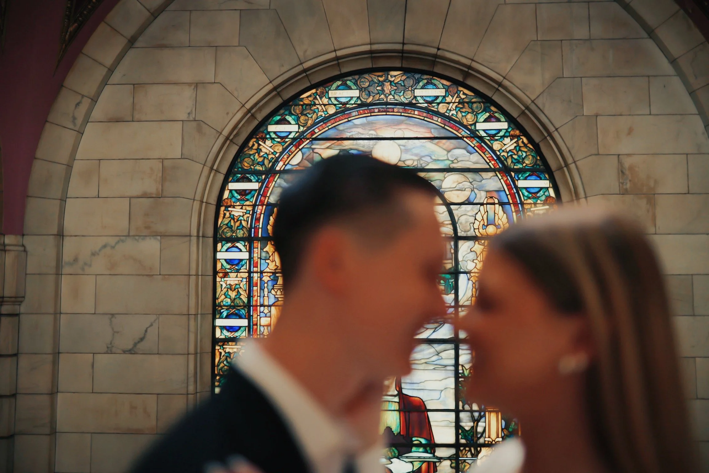 Blurred couple smiling and about to kiss in front of a stained glass window with biblical imagery in a church.