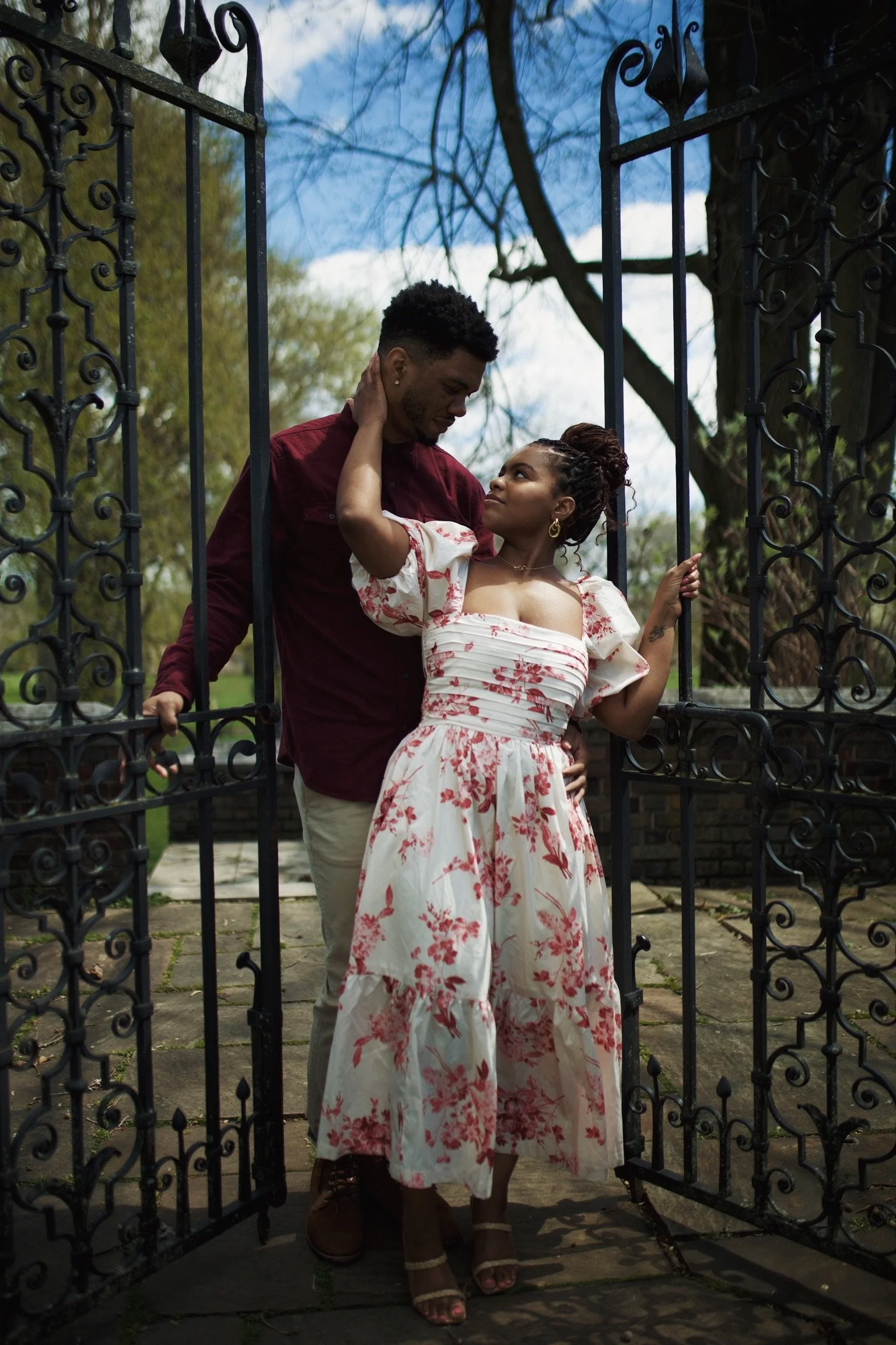 A couple stands together in a garden gate, with the man gently touching the woman's face and she looking up at him affectionately.