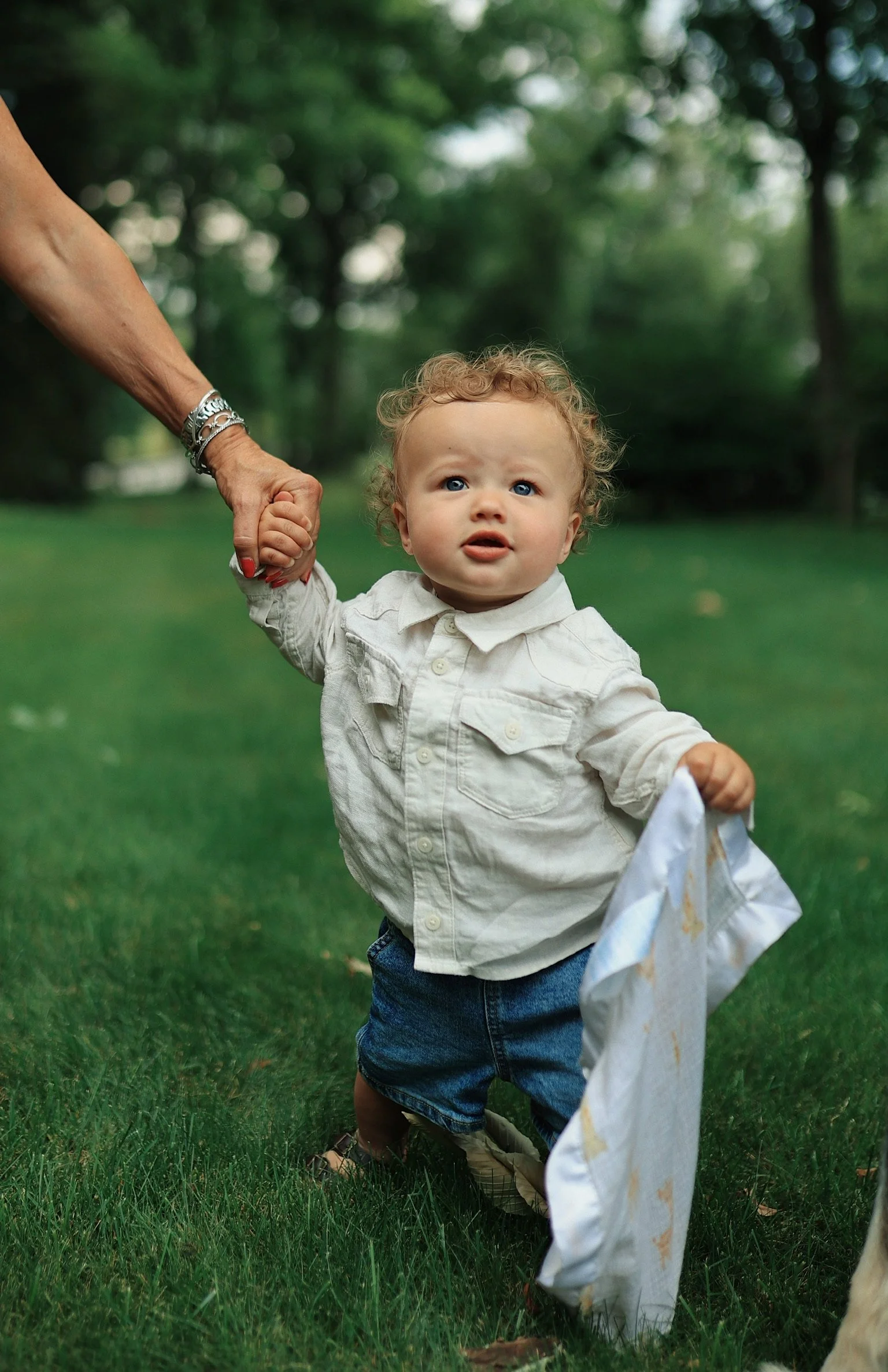 A young child with curly hair and blue eyes holding a woman's hand while walking on a grassy area outdoors.