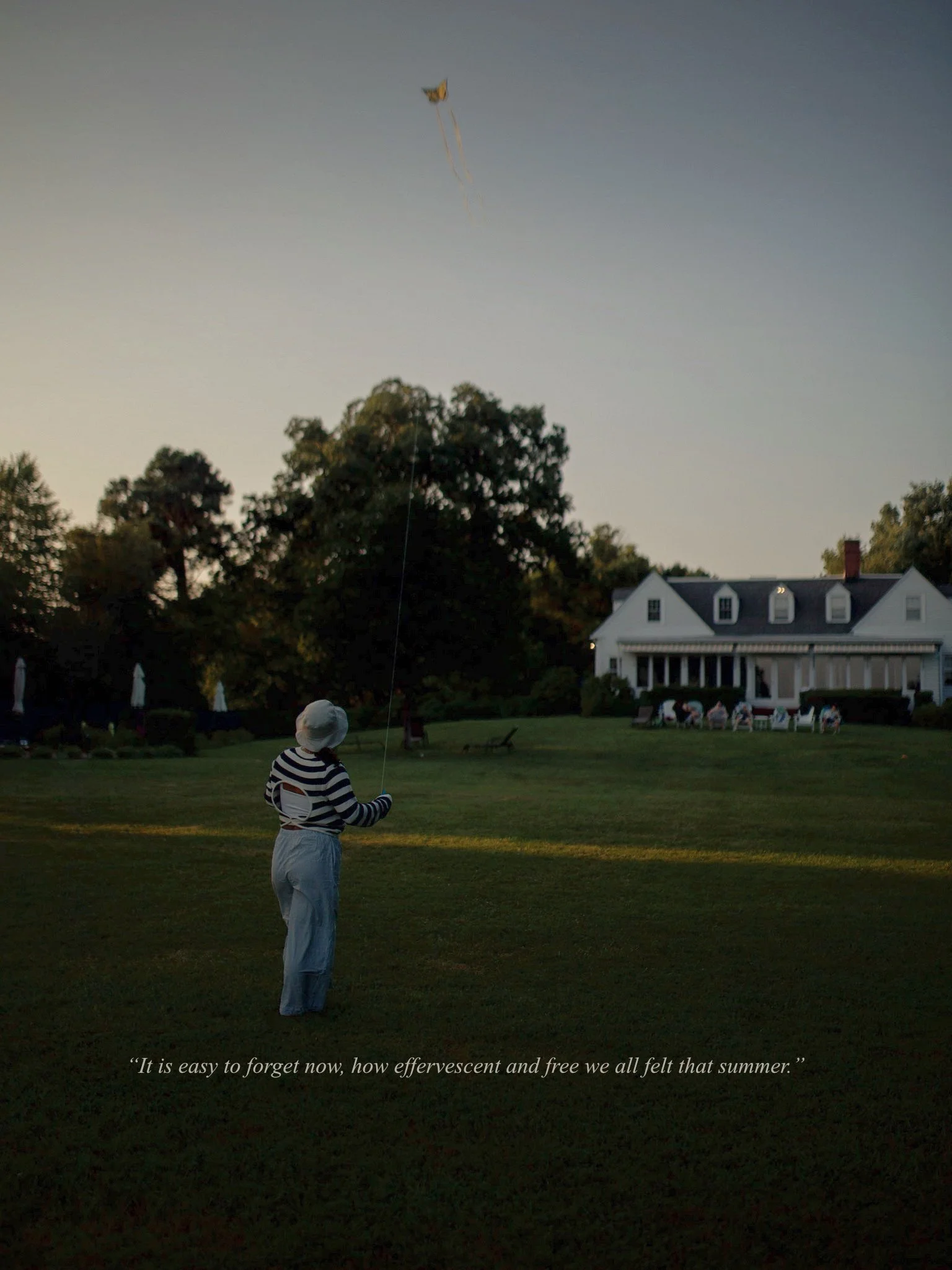 A person flying a kite in a large backyard at sunset with a big house in the background.
