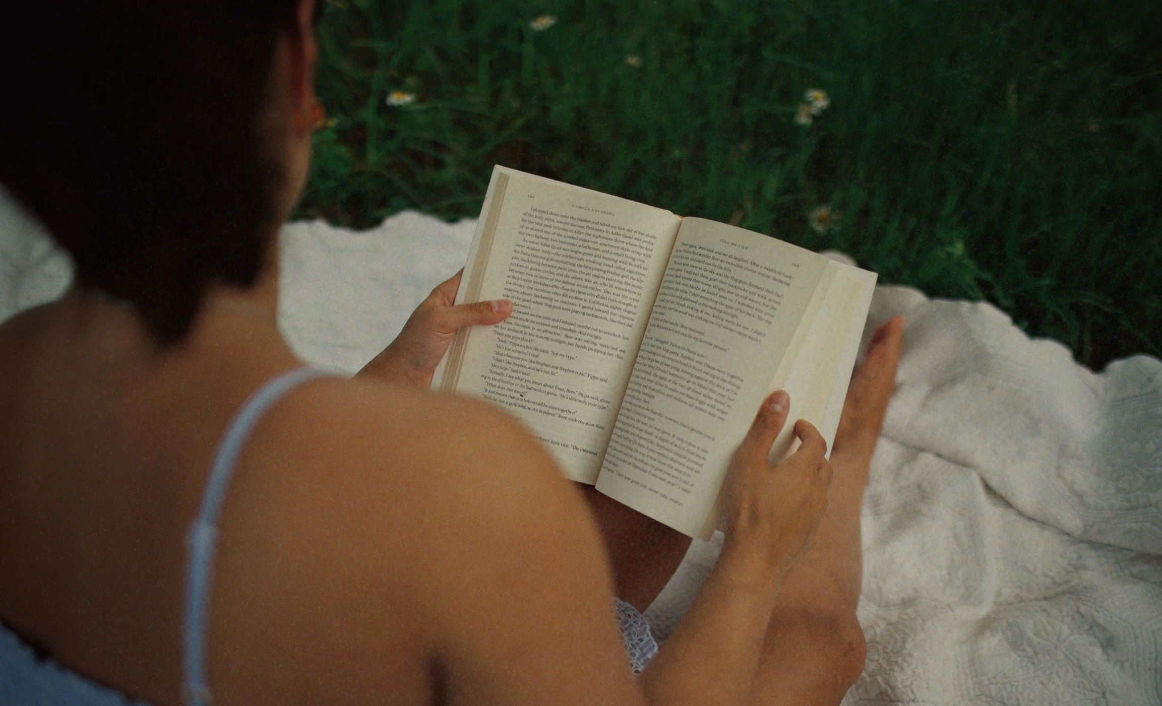 A person with a white strap top reading a book outdoors on a white surface with green plants in the background.
