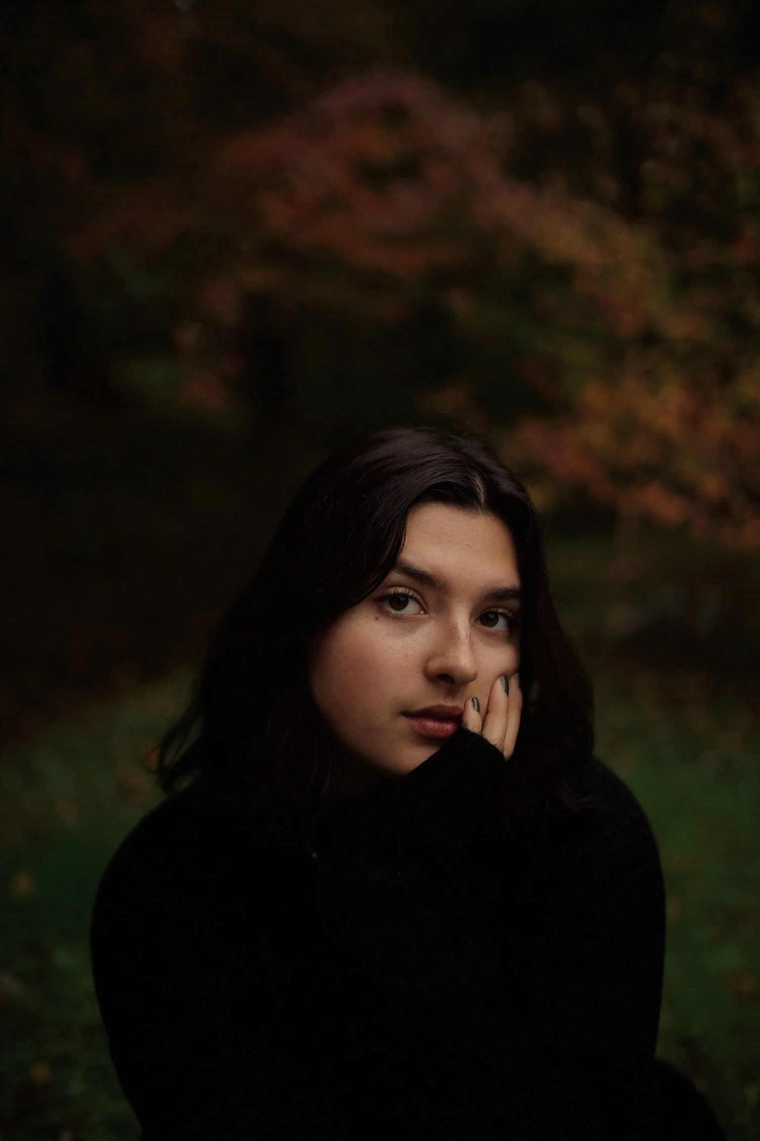 A young woman with dark hair and light skin, wearing a black sweater, stands outdoors in a dimly lit environment with autumnal trees in the background.