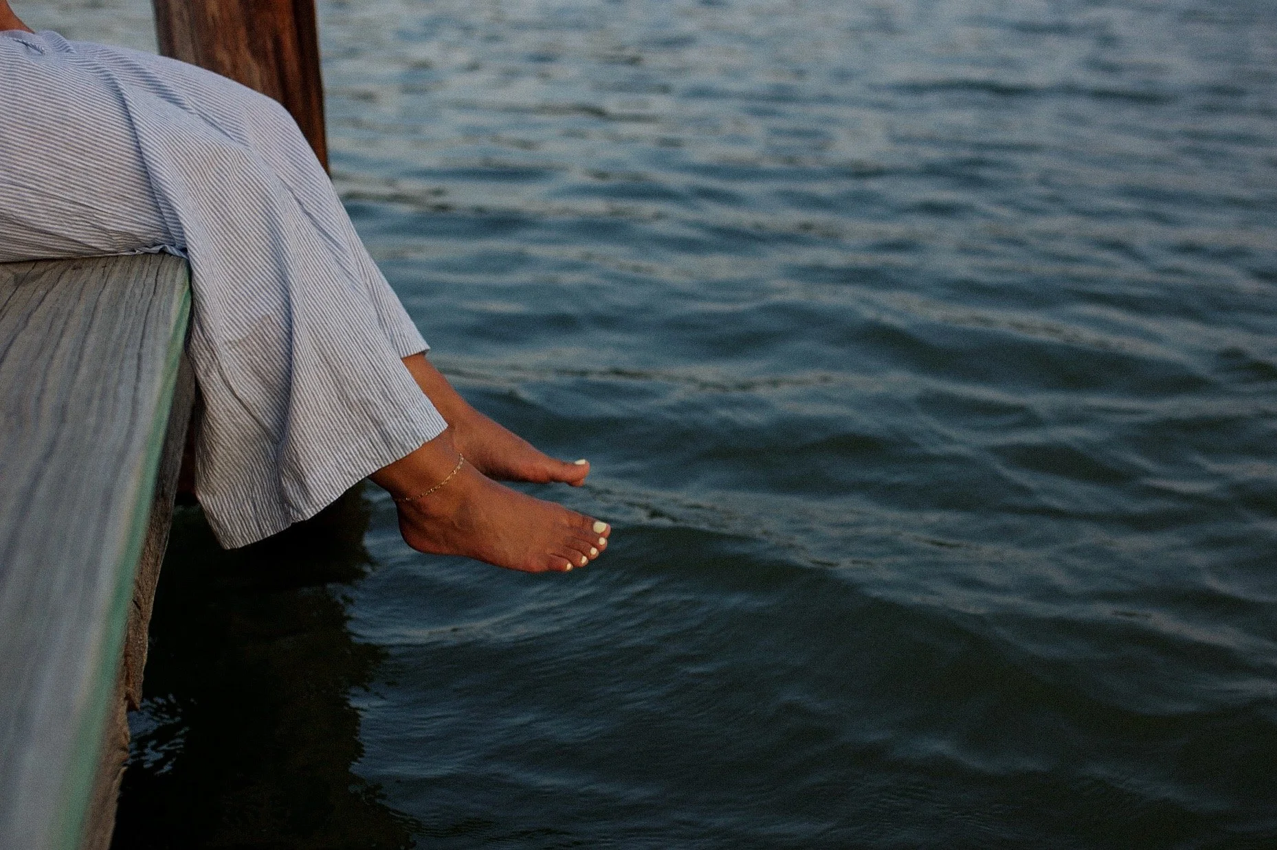 A person sitting on a wooden dock with their legs and feet hanging over the water, wearing striped pants, an anklet, and painted toenails.