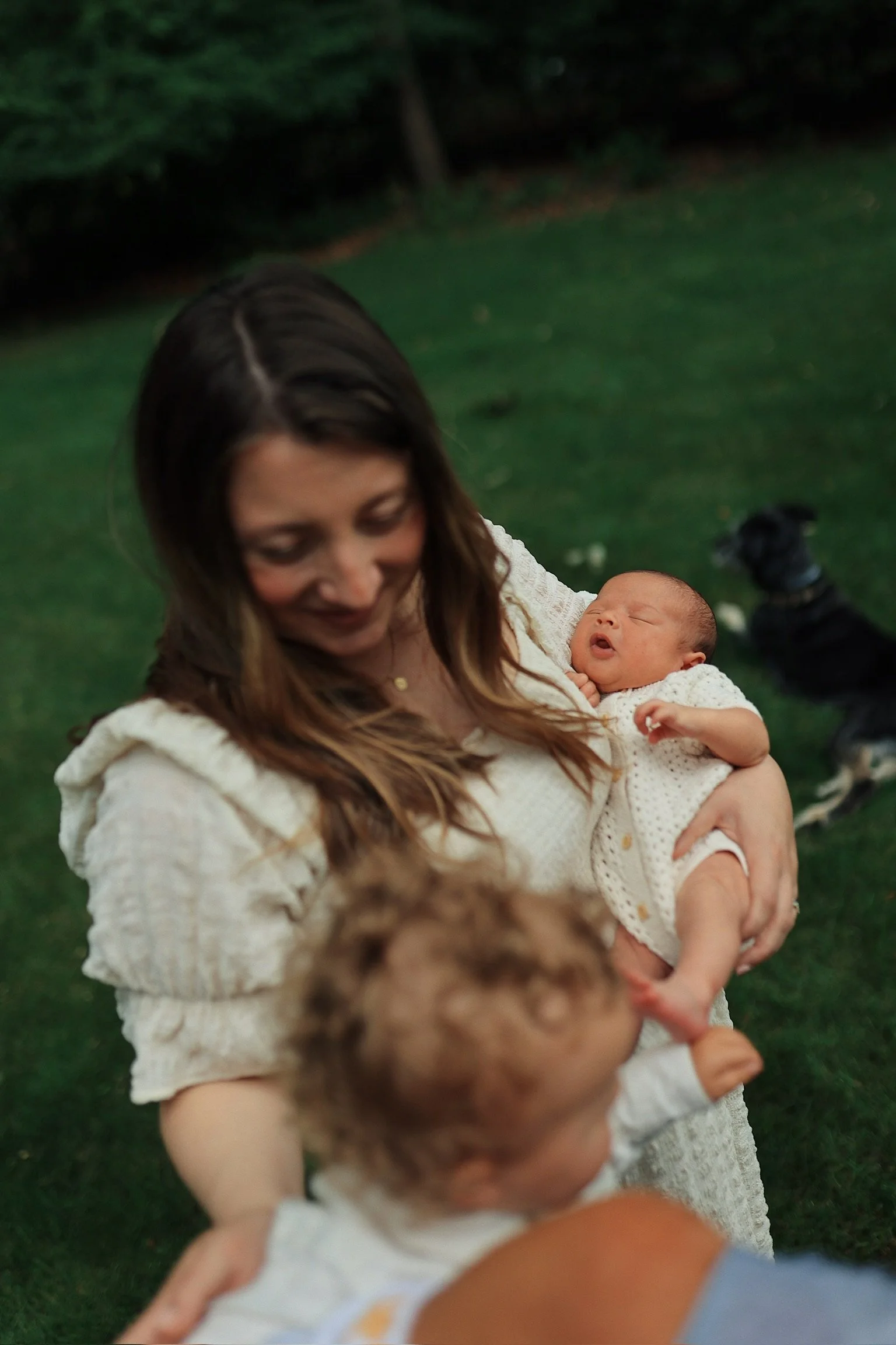 A woman holding a baby outdoors on a grassy area with trees in the background. Another person with curly hair and a dog in the background.