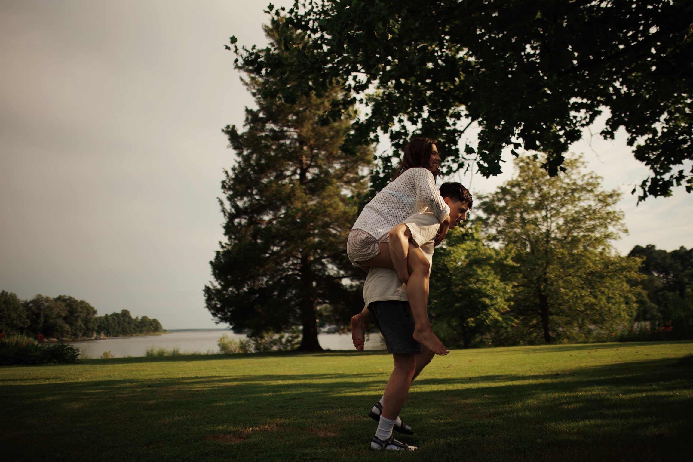 A man carrying a woman on his back in a park with trees and a body of water in the background.