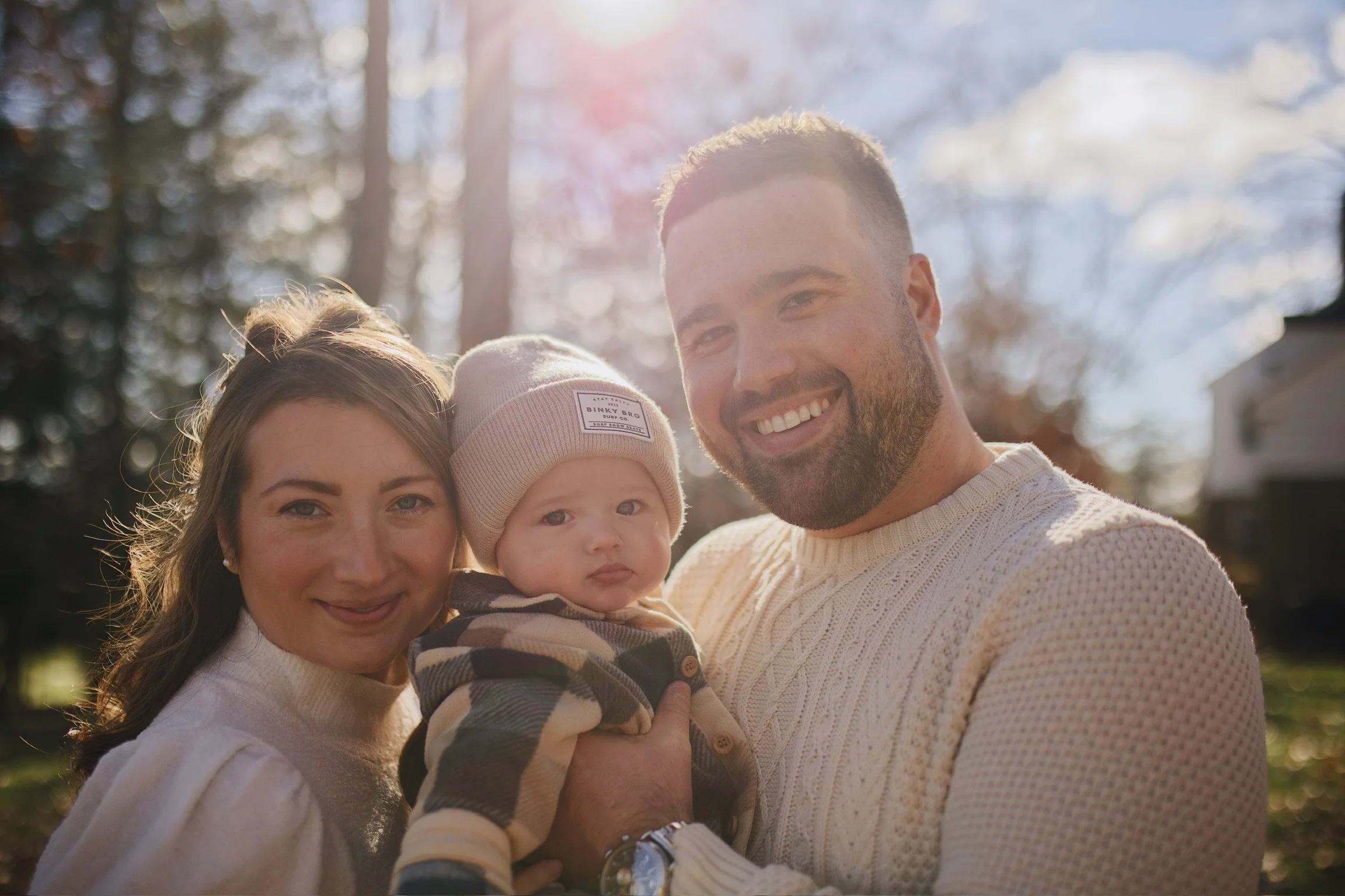 A smiling family of three with a woman, a man, and a baby outdoors in a park with trees in the background, sunlight shining through, during fall or winter.