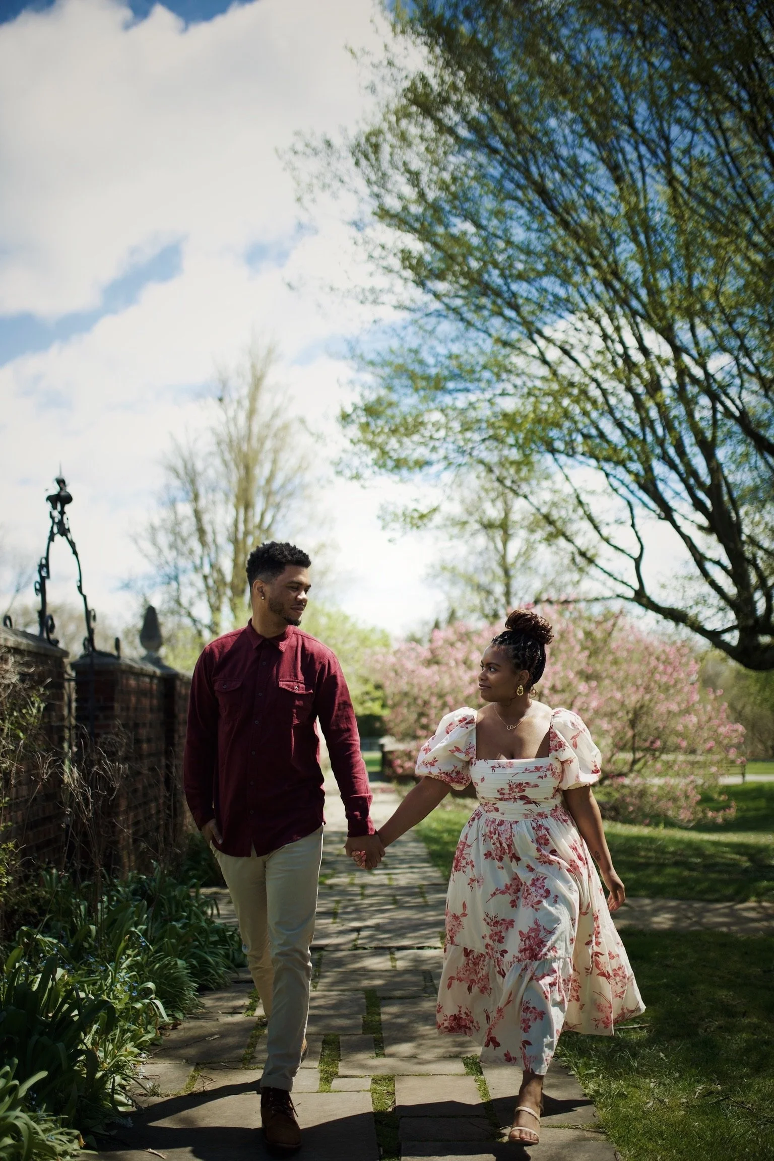 A couple holding hands and walking along a garden path on a sunny spring day, with blooming trees and a clear sky in the background.