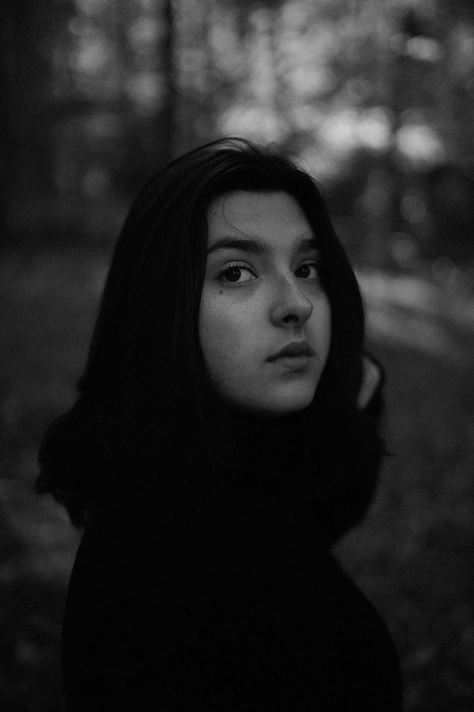 A black and white photo of a young woman with dark hair, looking at the camera, outdoors with blurred trees in the background.