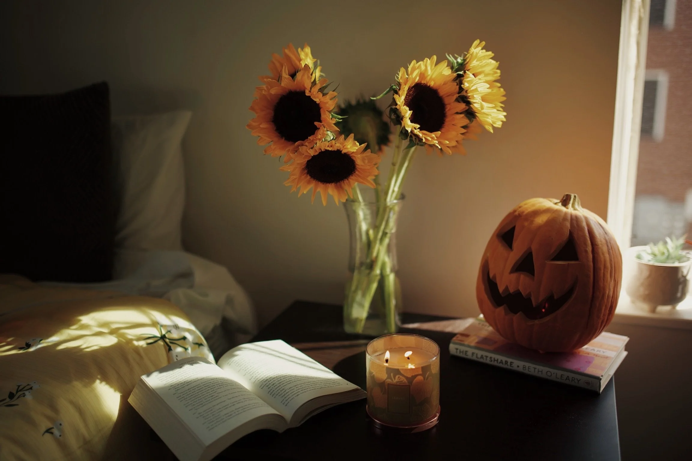 A cozy room corner with a sunflower bouquet in a glass vase on a nightstand, a lit candle, an open book, a pumpkin carved as a jack-o'-lantern, and a small stack of books near a window with soft natural light.