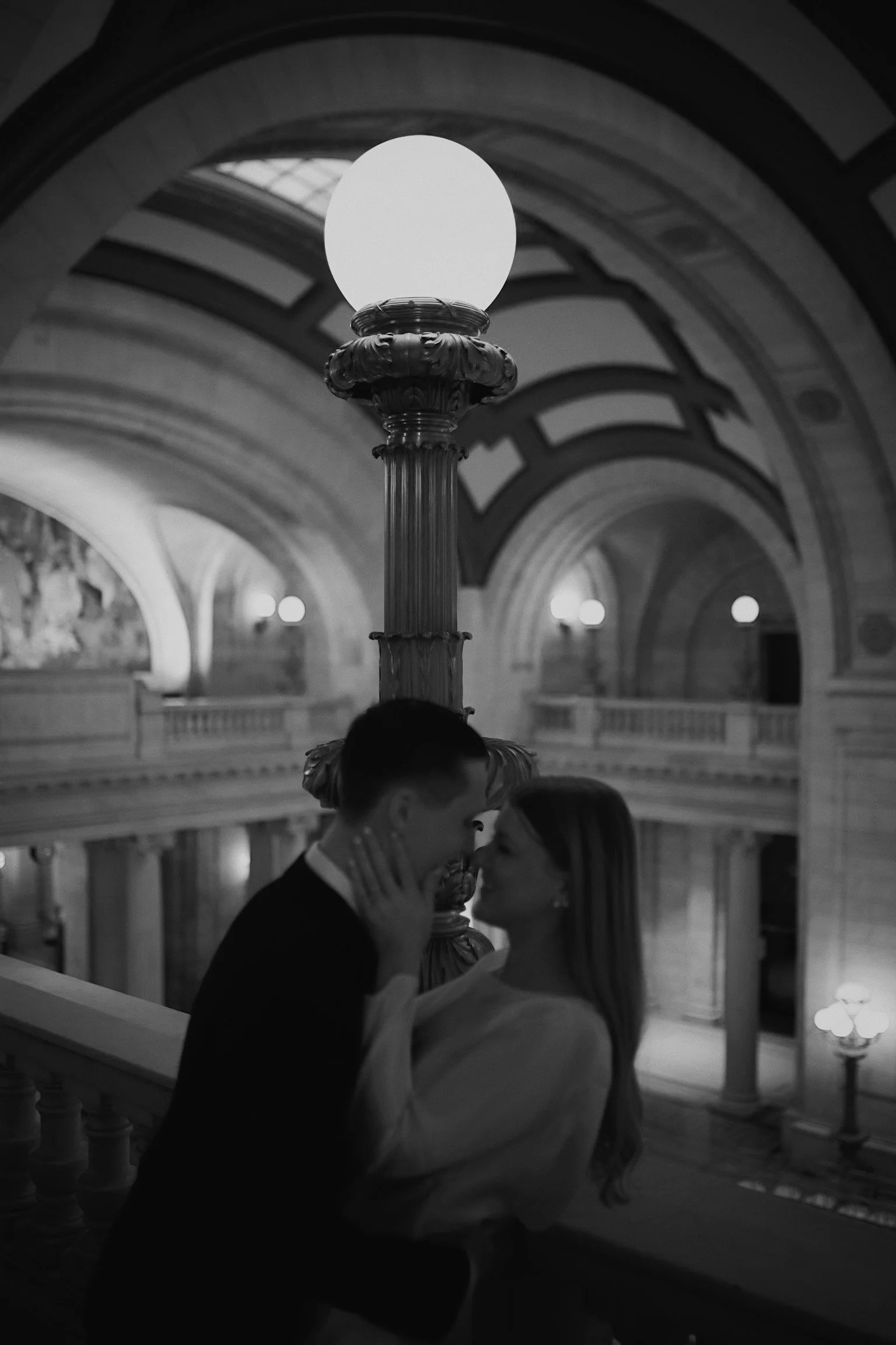 A couple, a man and a woman, sharing an intimate moment on a staircase in a grand, ornate building with arched ceilings and classical architecture, illuminated by a large vintage lamp.