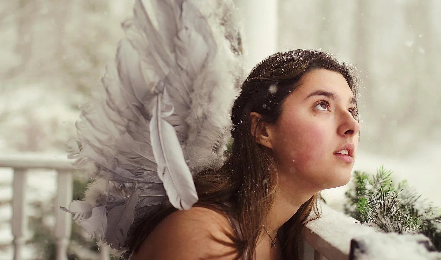 Young woman with angel wings looking out a window on a snowy day.
