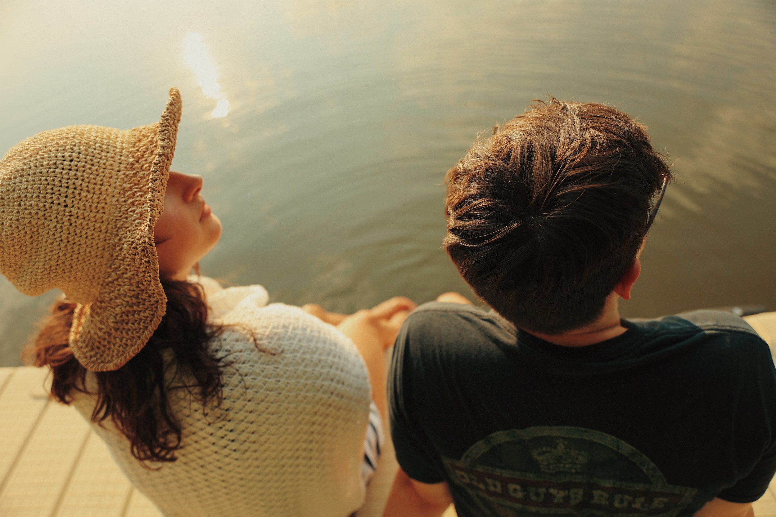 A woman and a man sitting on a dock by a lake, seen from behind, relaxing and enjoying the peaceful water.