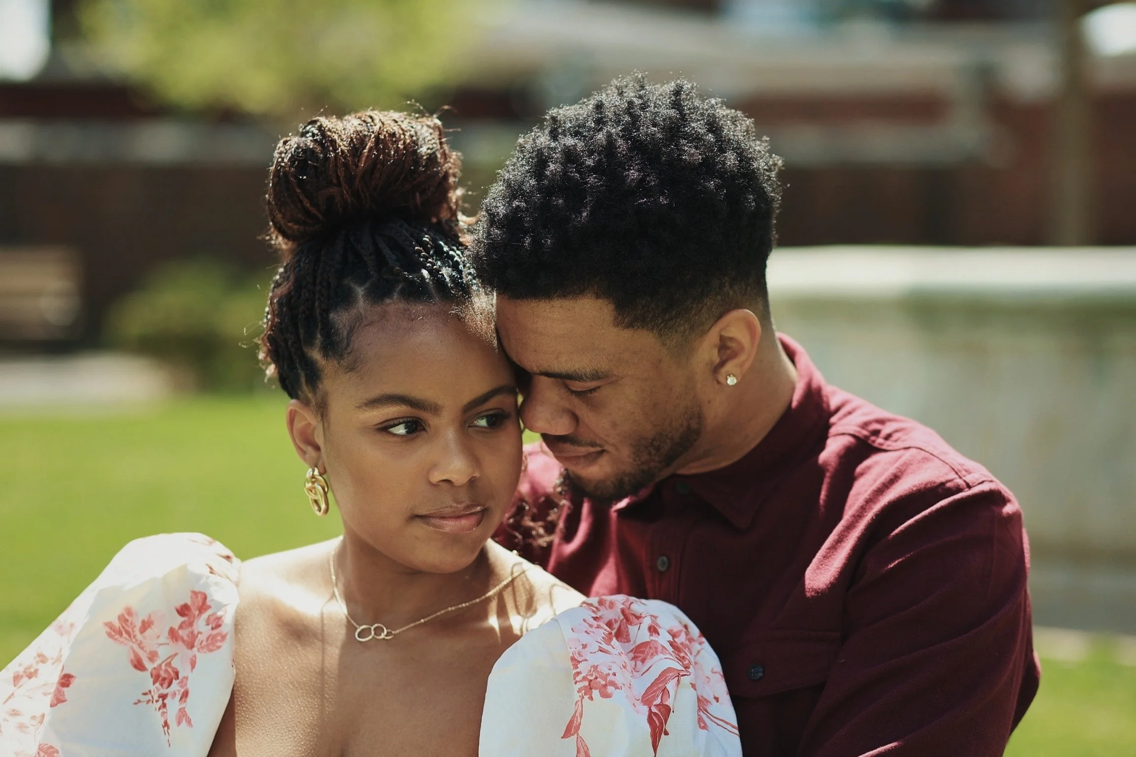 A couple facing each other outdoors, with the man resting his forehead against the woman’s forehead, both with serious expressions.