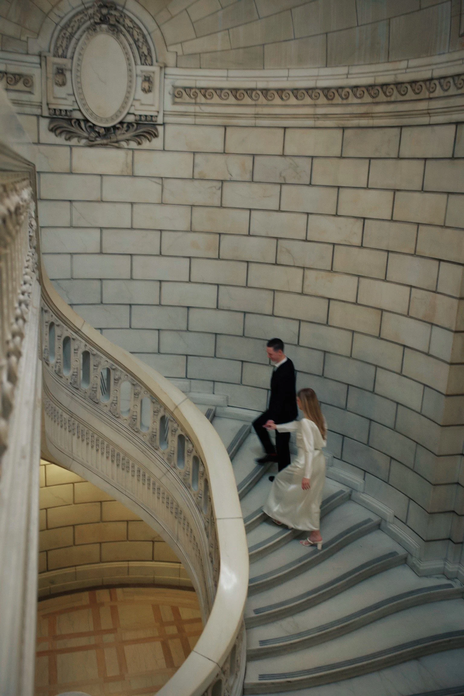 A man in a black suit and a woman in a white dress walking down a curved marble staircase inside a building with stone walls.