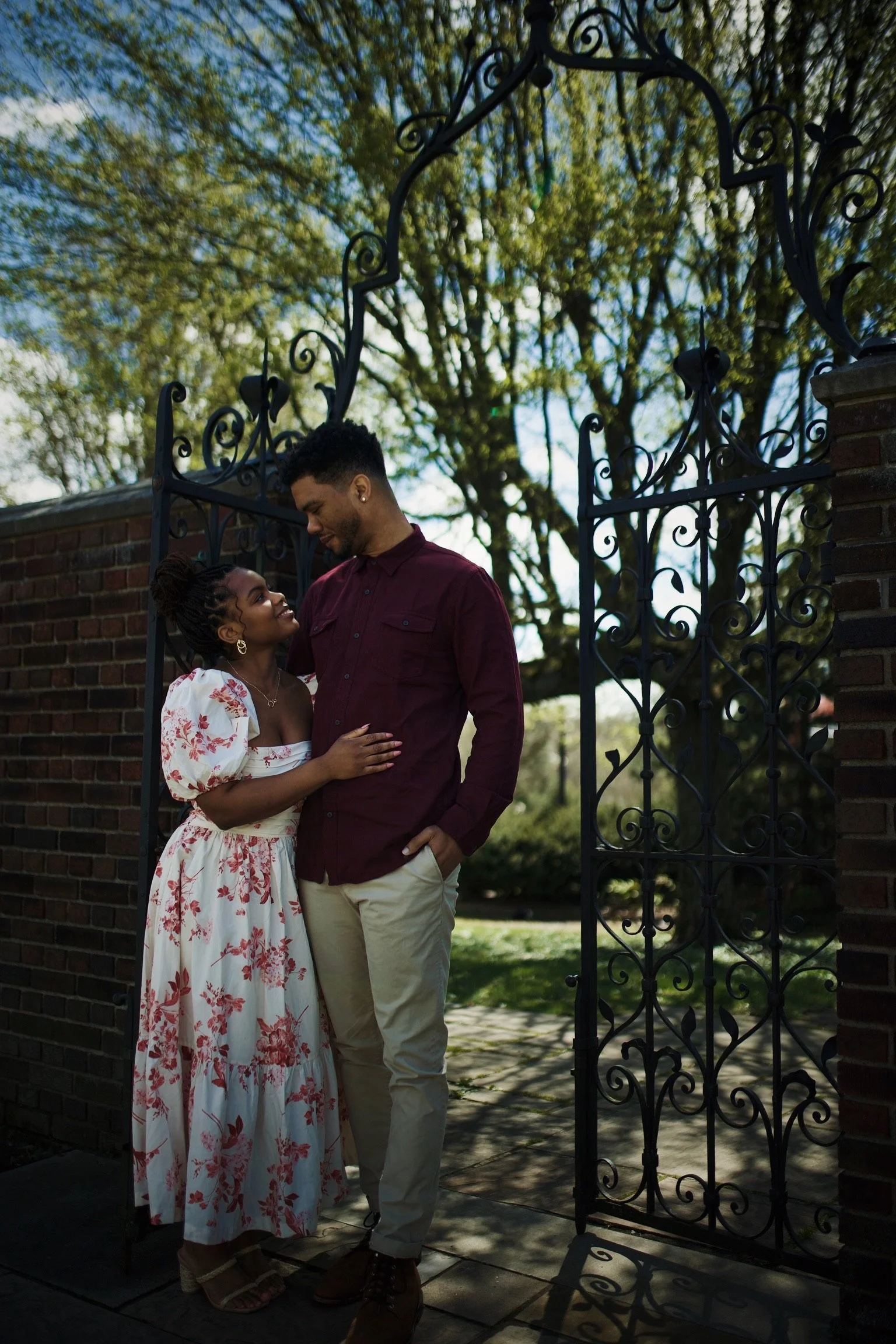 A couple standing near a decorative iron gate, embracing and smiling at each other outdoors during daytime, with trees and blue sky in background.