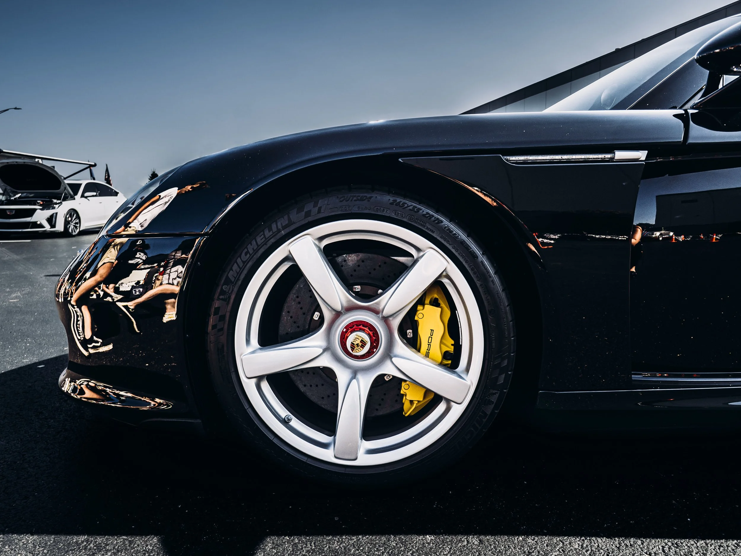 The image shows a close-up of a black Porsche sports car, focusing on its front left wheel. The wheel features a shiny silver alloy rim with a red and yellow Porsche emblem at its center. The brake caliper is painted bright yellow and has the “Porsch