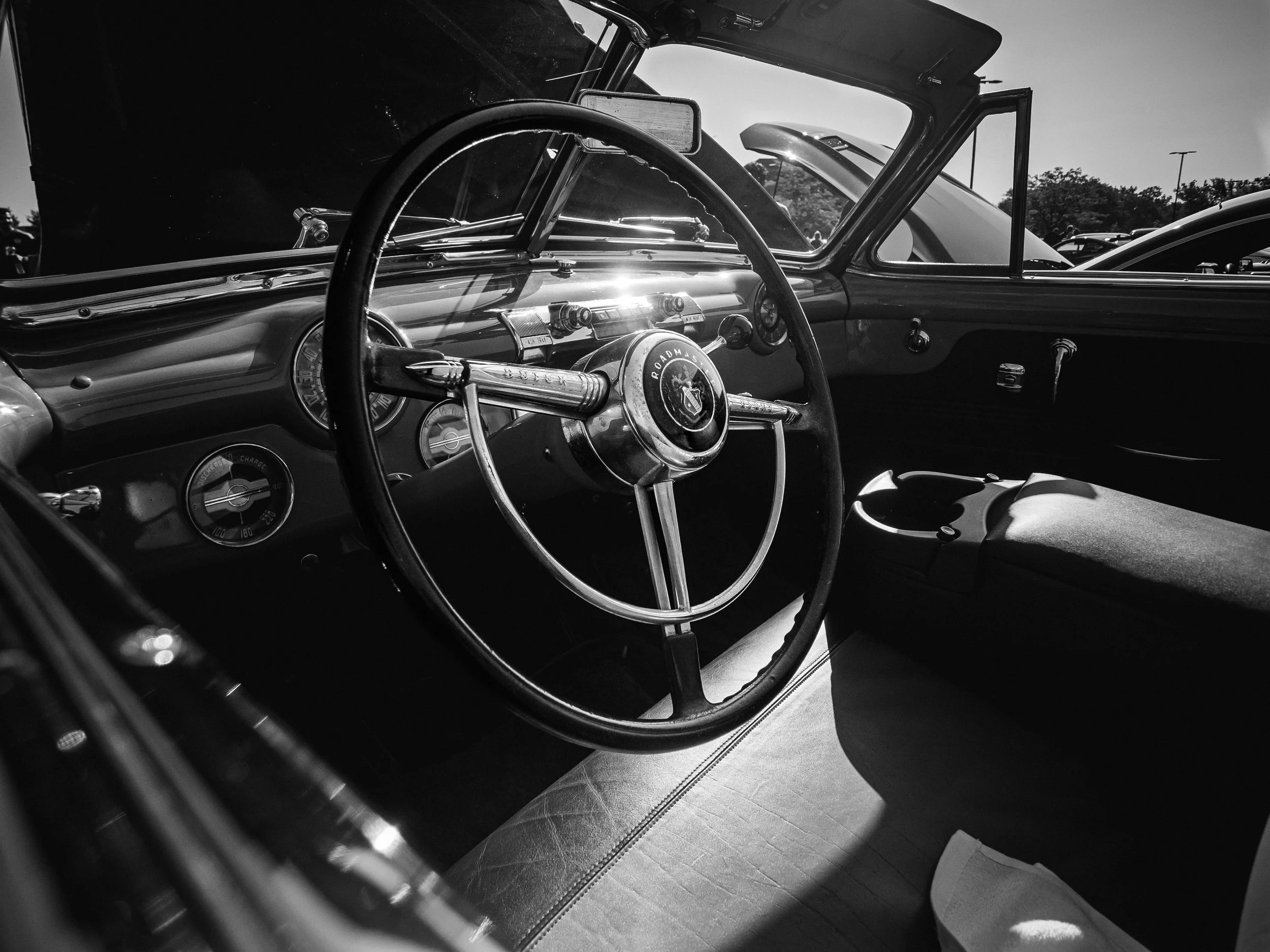 The black and white photograph captures the interior of a vintage Buick Roadmaster, focusing on the driver's side. The image showcases a large, polished steering wheel with a prominent horn ring and "Roadmaster" insignia in the center. Behind the ste