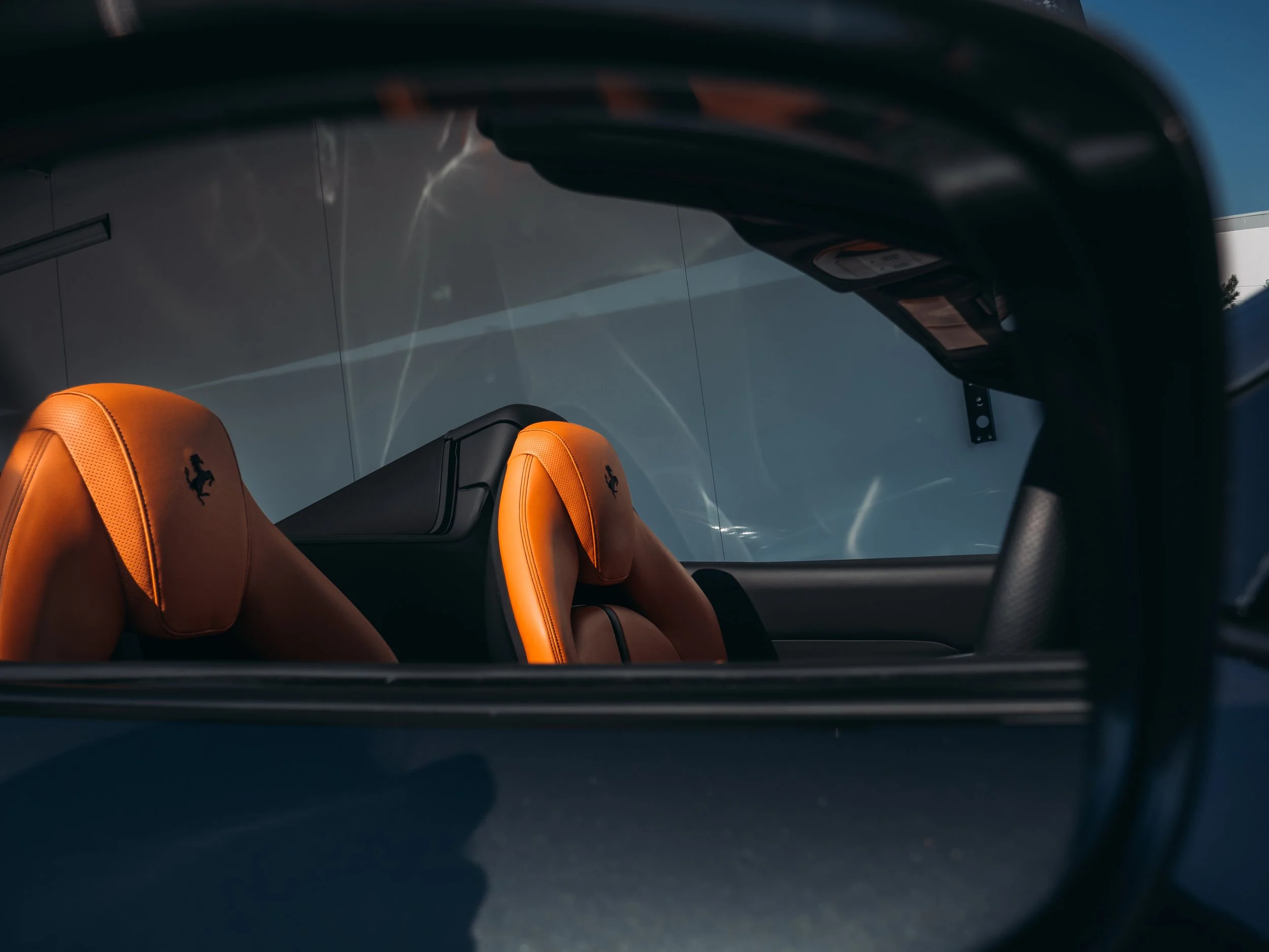 The image is a close-up view focusing on the side mirror of a Ferrari, capturing the reflection of the car's interior seats. The side mirror frame occupies the foreground, featuring sleek and glossy black contours. In the reflection, two sporty, oran