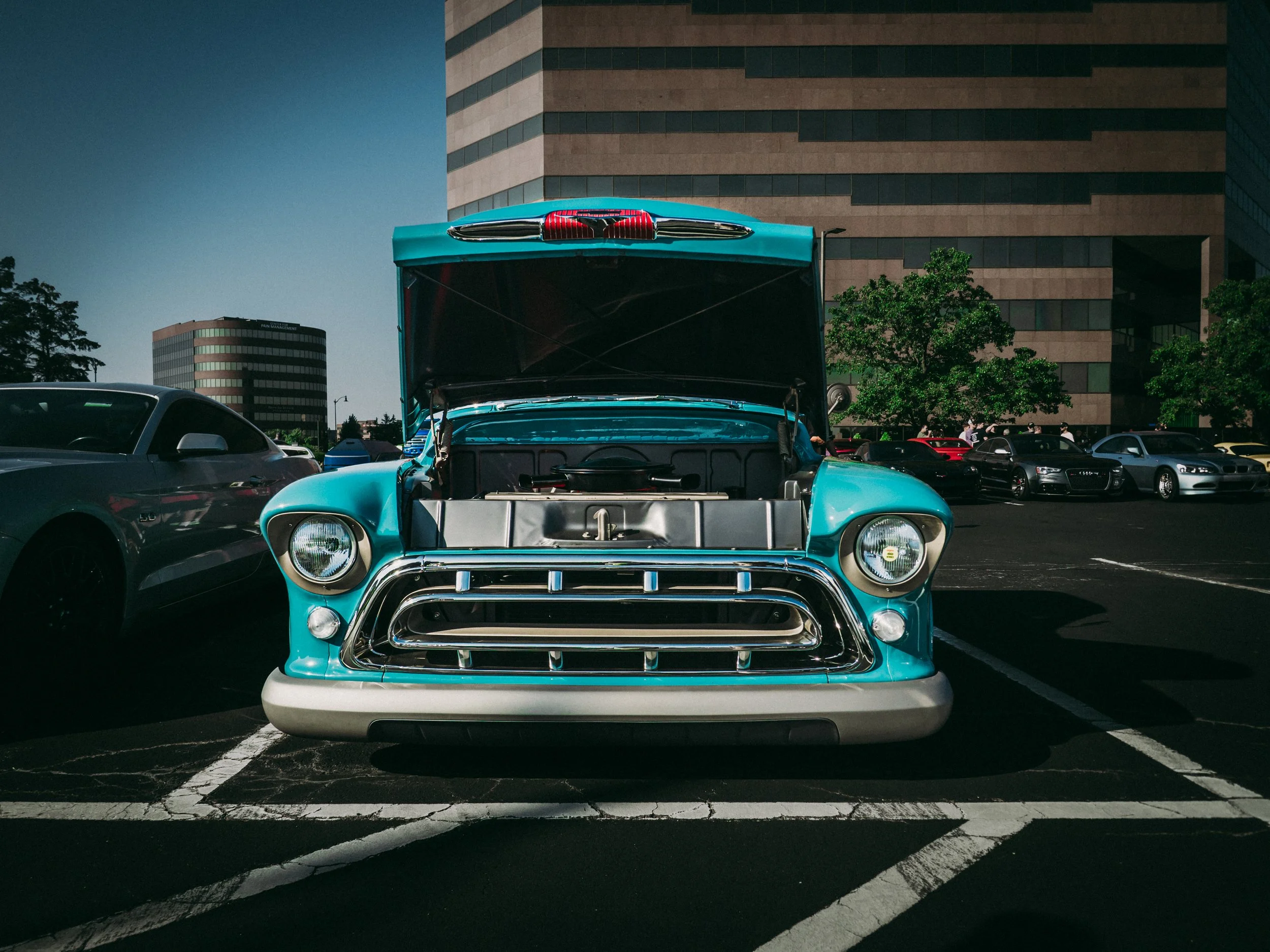 The image shows a classic turquoise Chevrolet truck with its hood open, parked in a lot against the backdrop of modern office buildings. The front view prominently features large, round headlights and a chrome grille with horizontal bars, typical of 