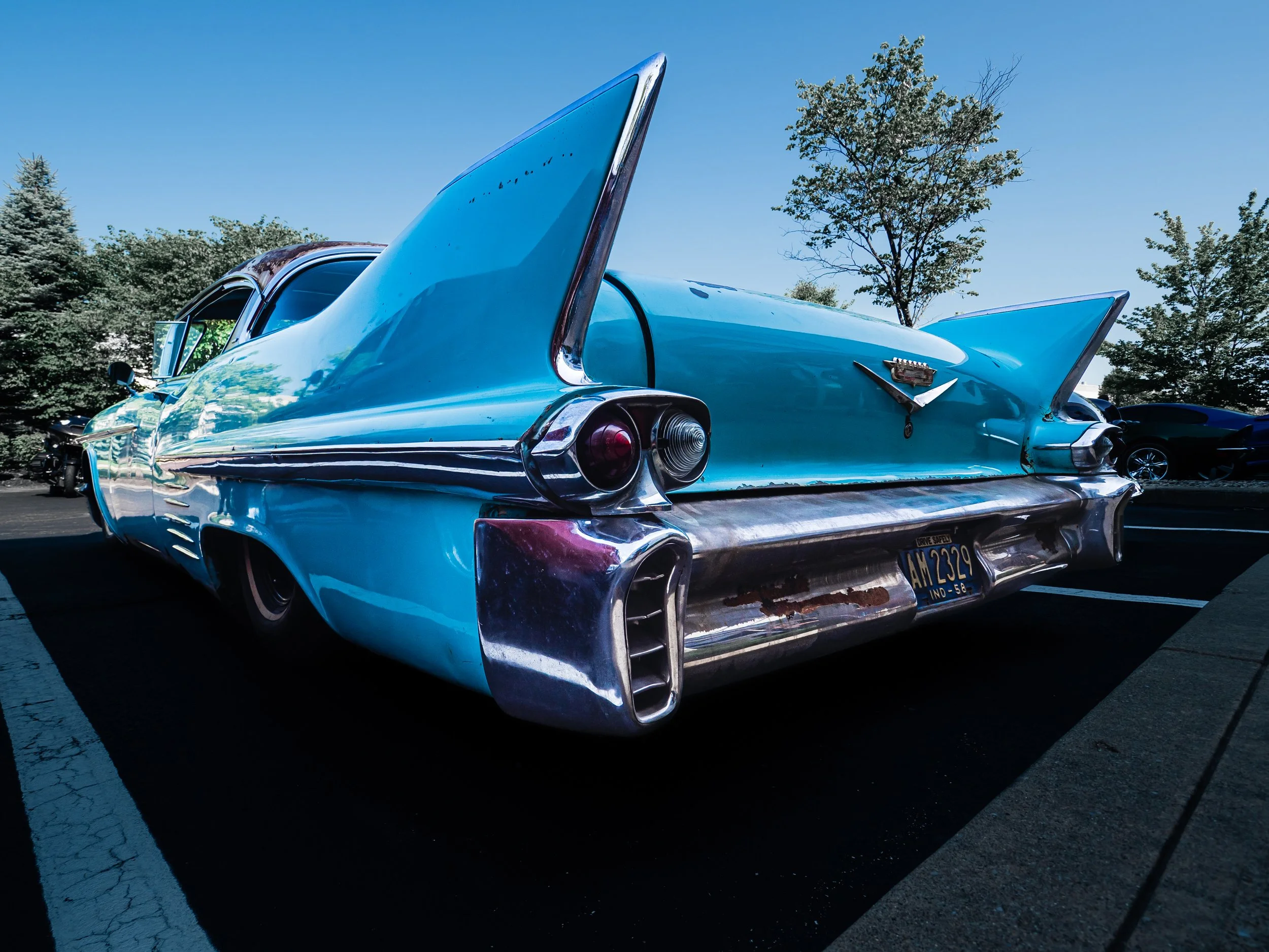 The image depicts the back left 45-degree angle of a vintage blue Cadillac parked in a lot. The car boasts prominent tailfins, typical of classic models, each containing a circular red taillight. The rear bumper is wide and chrome-plated, reflecting 