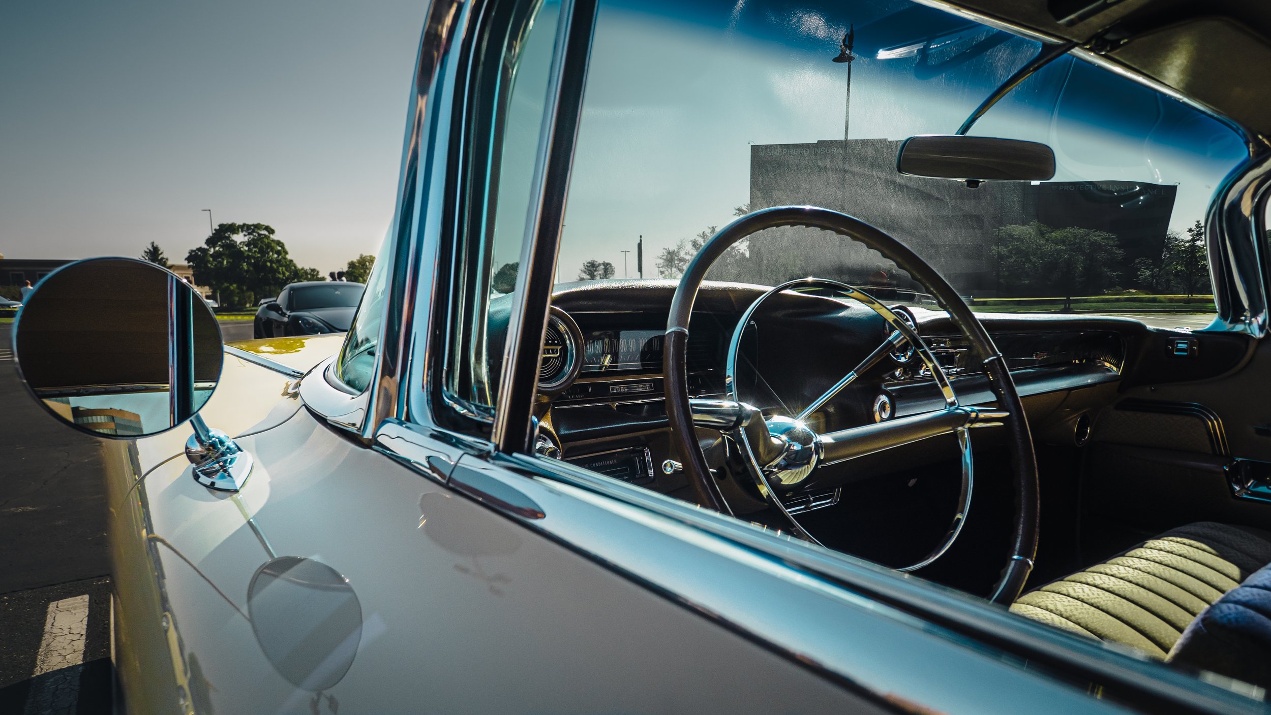 The image depicts a close-up view of the interior of a vintage Cadillac from an exterior perspective. The camera angle focuses through the driver's side window, capturing the steering wheel, dashboard, and part of the driver's seat. The Cadillac's ex