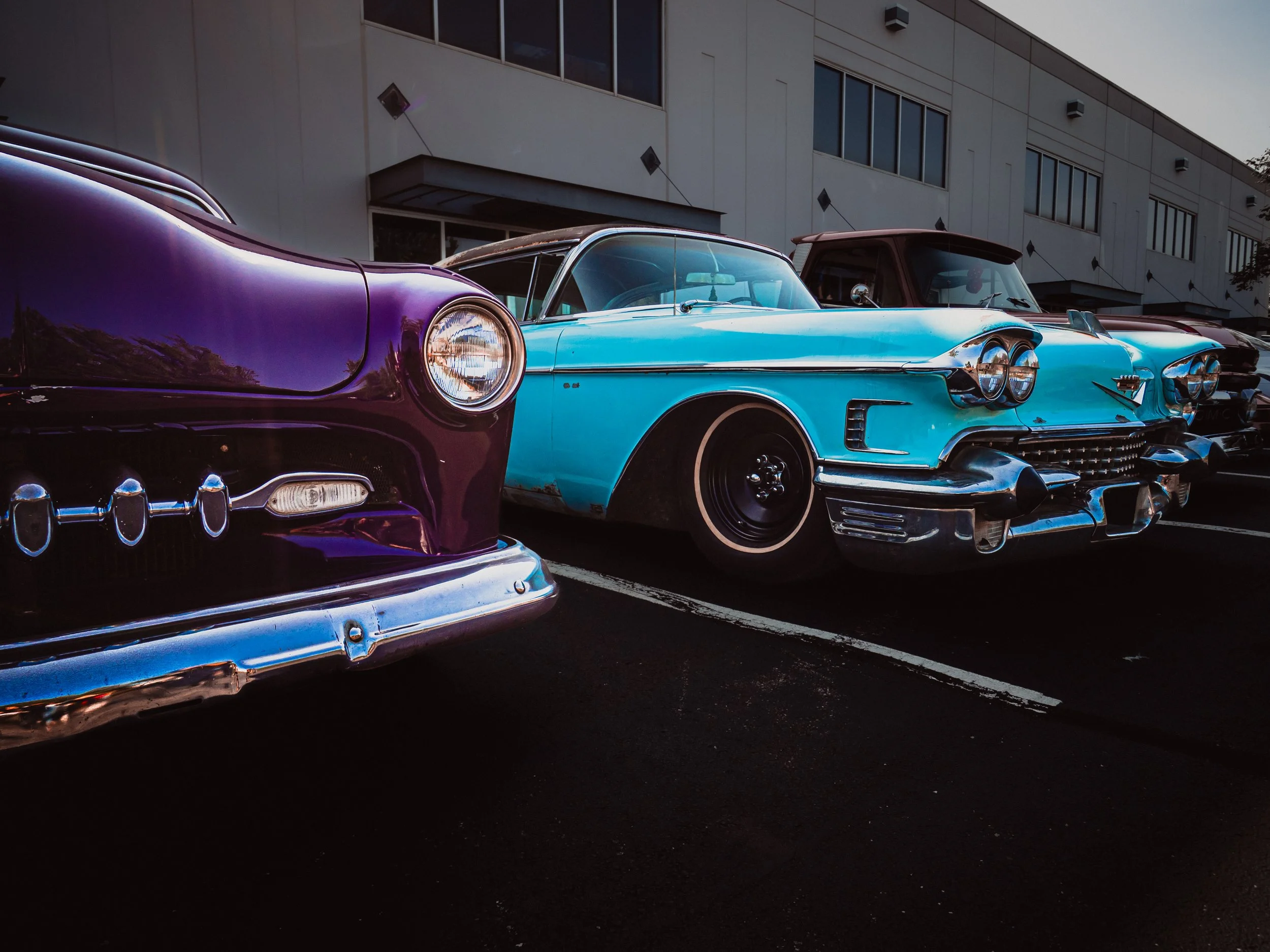 The image depicts a line-up of vintage cars parked outside a modern industrial-looking building with large windows. The focus of the image is on two classic cars— a Ford on the left and a Cadillac on the right. The Ford is a deep, glossy purple color