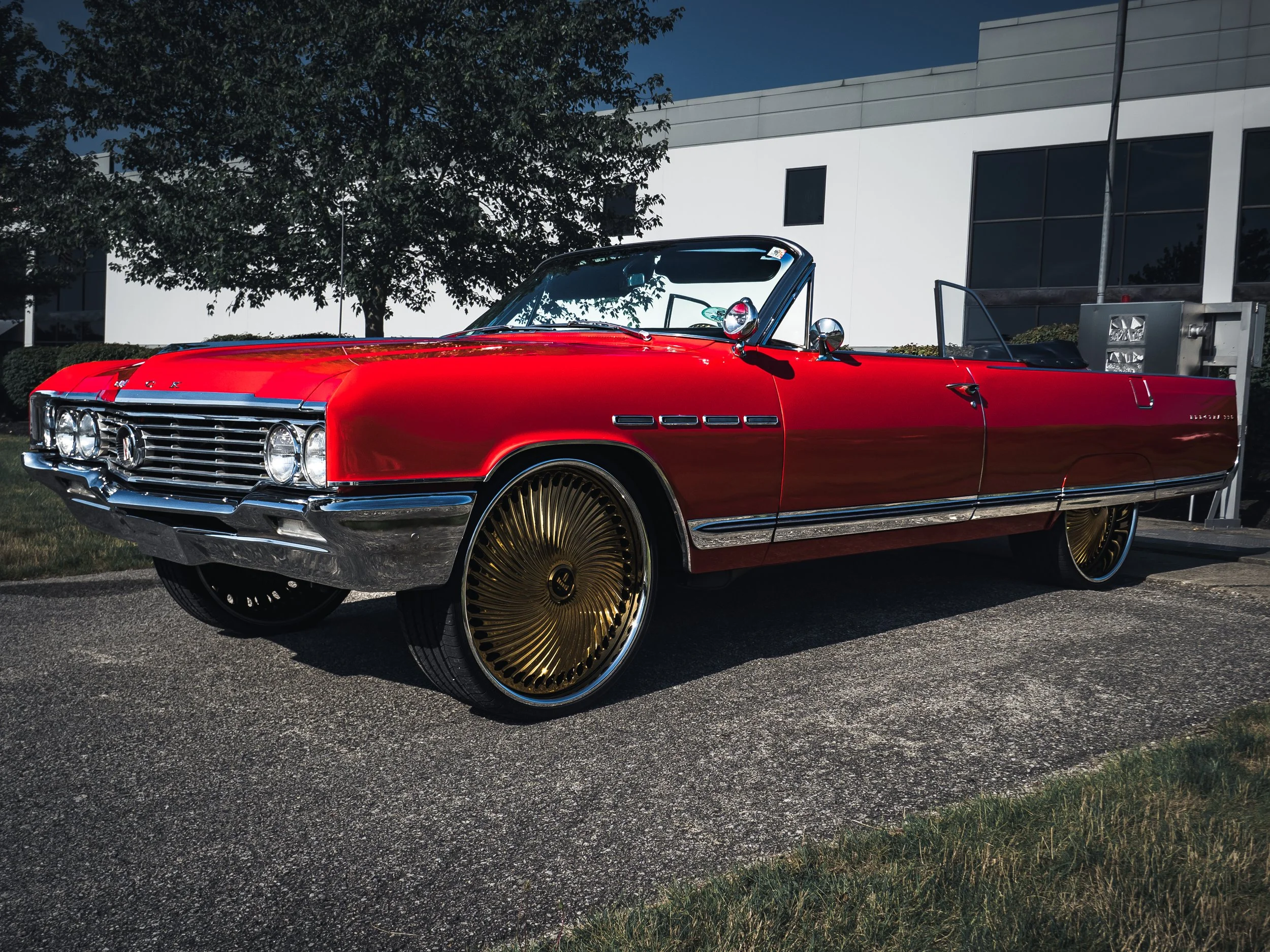 The image features a classic red Buick convertible car viewed from the front left side at a 45-degree angle. The car has a shiny chrome grille with horizontal slats and four round headlights, two on each side. The body of the vehicle is sleek and pol
