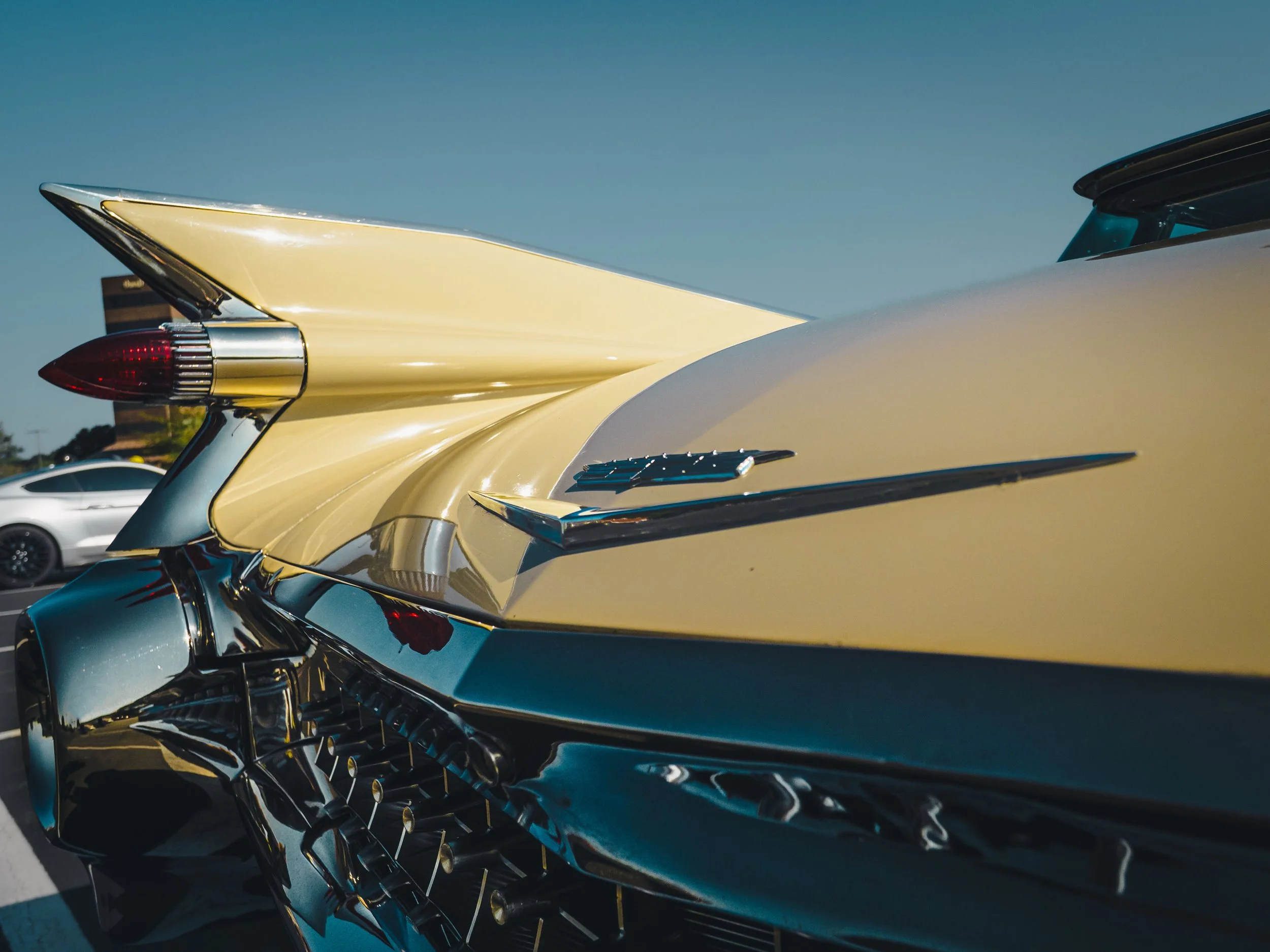 The image depicts a close-up view of the back right end of a classic Cadillac car. The Cadillac is painted in a soft yellow color and features prominent, sleek, and shiny chrome detailing. The tail fin, a notable characteristic of classic Cadillacs, 