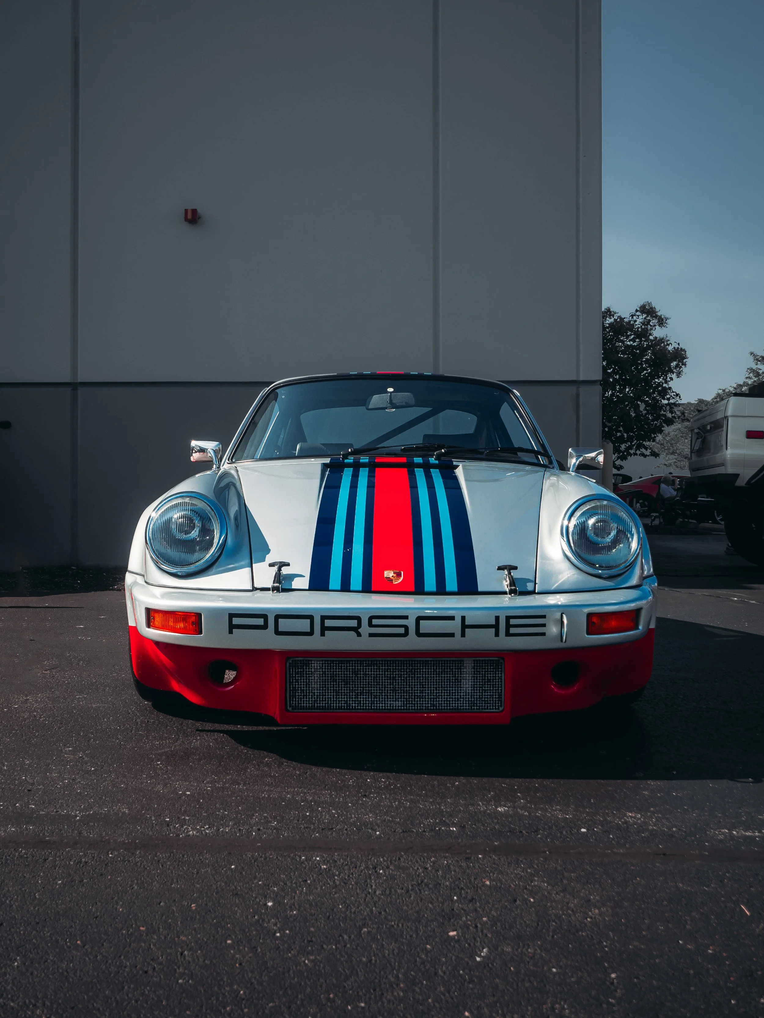 The image features a straight-on view of a classic Porsche car, parked against a plain, light-colored wall with minimal elements in the background. The car is predominantly white and adorned with distinctive vertical racing stripes in blue and red. T