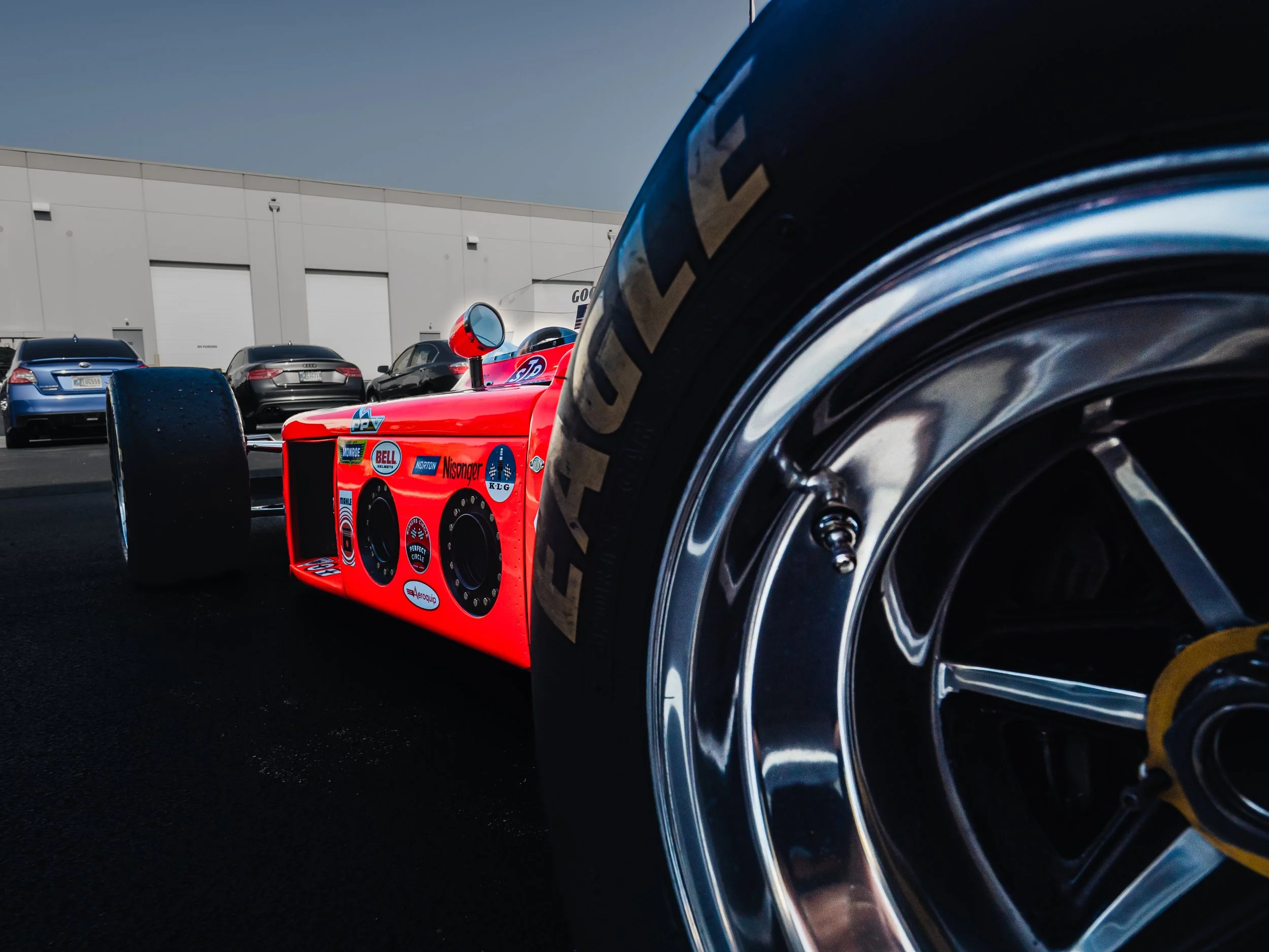 The image shows the back left side of a bright orange Indy 500 race car. The main focus is on the large, glossy tire in the foreground, with the word "EAGLE" visibly printed in gold on its sidewall. Visible behind the tire is the orange body of the r