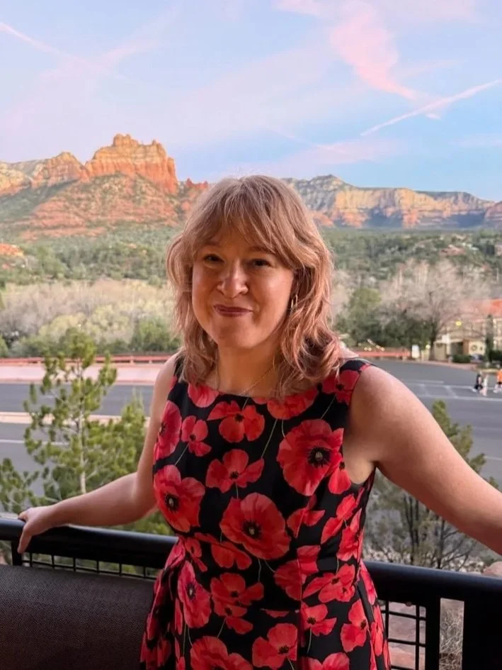 redheaded middle aged white woman standing in front of Sedona, Arizona mountains with a red poppy dress. Strategic coach Rachel Strivelli smiles at the camera in the sunset.